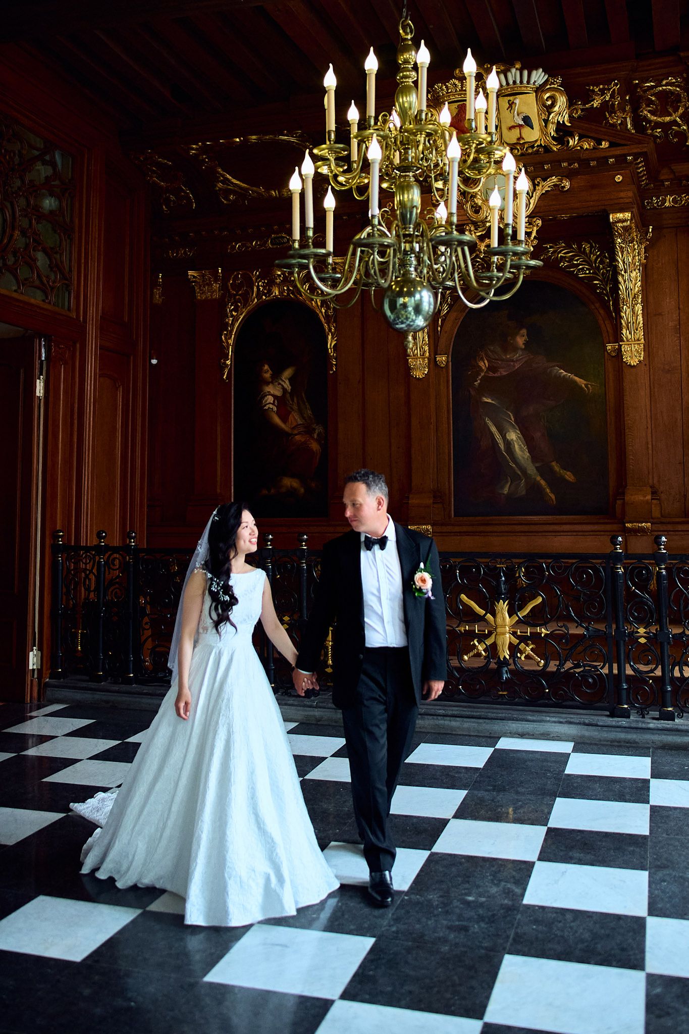 Couple walking hand in hand across a checkered ballroom floor beneath an ornate crystal chandelier.
