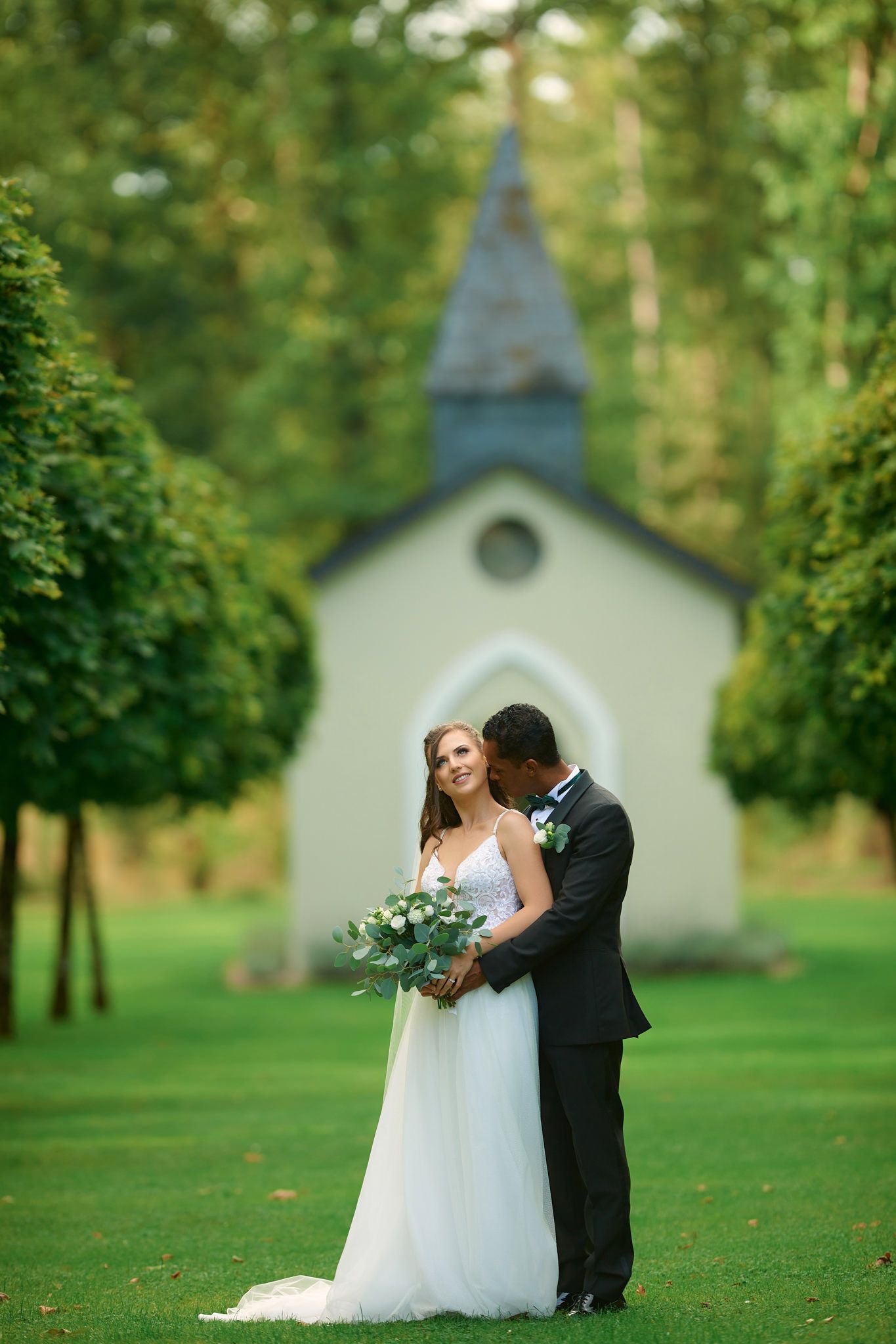 Romantic wedding portraits in front of a small chapel surrounded by lush green trees.