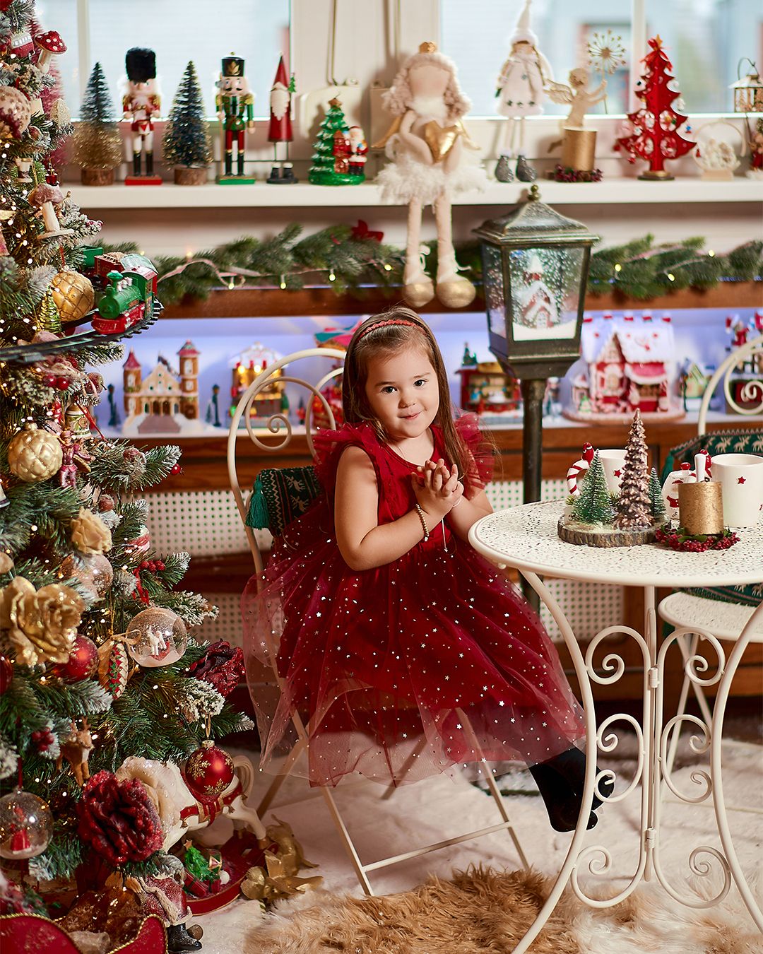 Young girl in red tulle dress poses beside decorated Christmas tree surrounded by festive holiday decorations and nutcrackers.