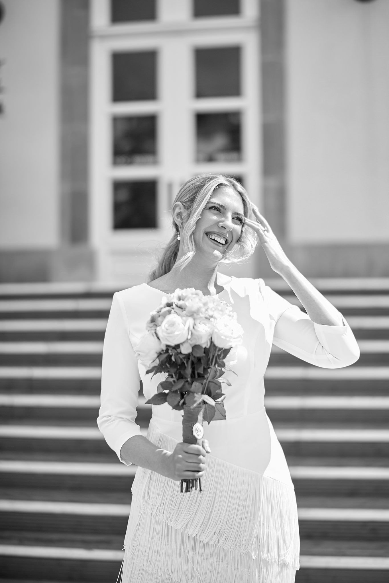 Joyful bride laughs while holding bouquet on outdoor steps in black and white photo.