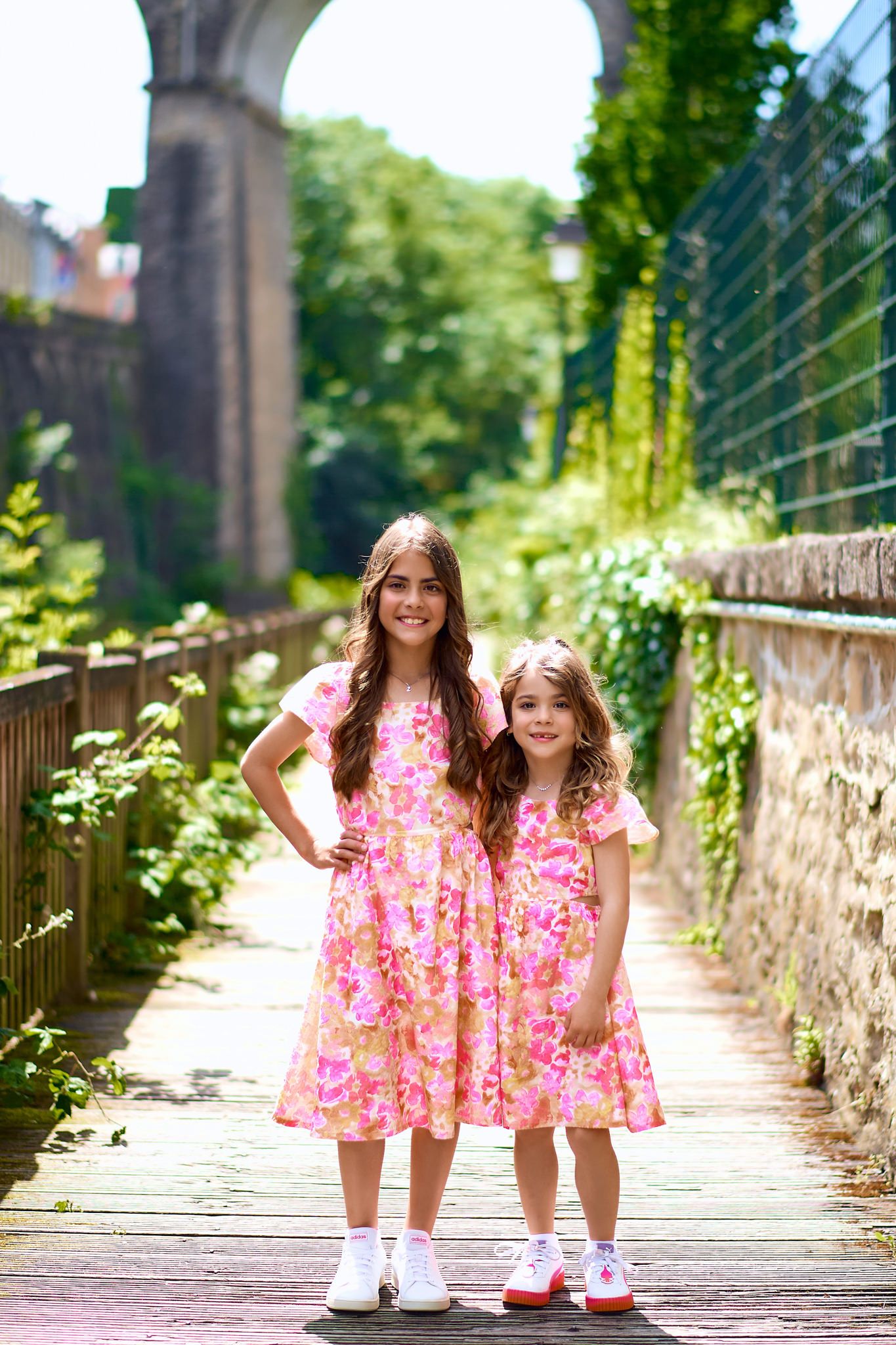 Two people in matching pink floral dresses pose on wooden walkway surrounded by greenery.