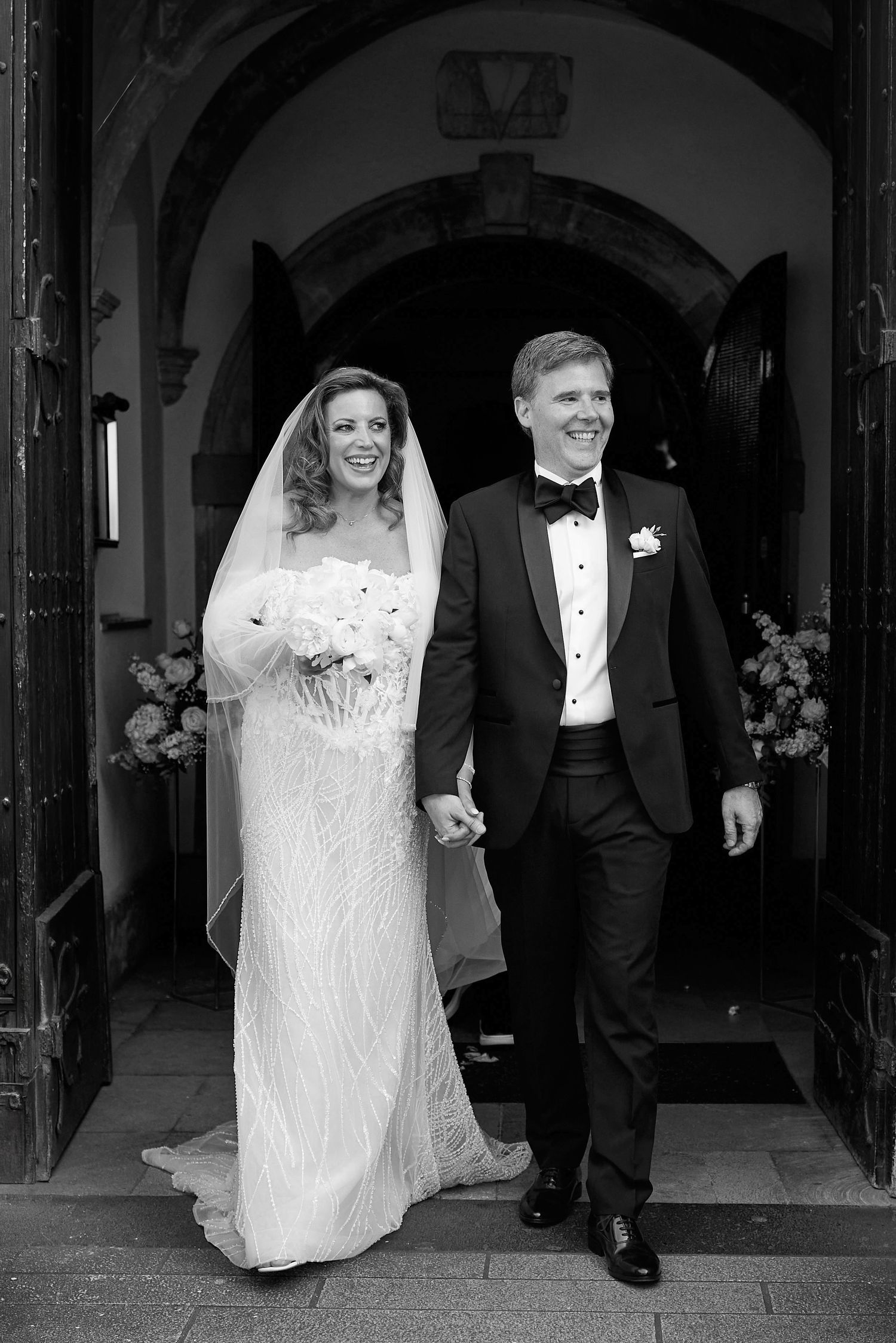 Black and white photo of a smiling bride and groom holding hands while exiting a church after their wedding ceremony.