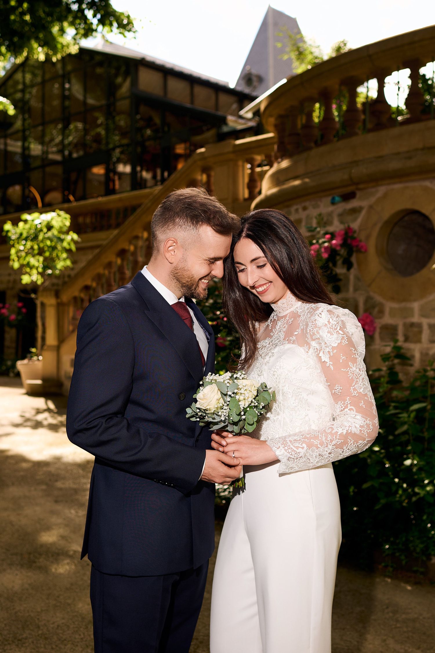 Smiling bride in lace gown holds bouquet with groom in navy suit outdoors at elegant stone venue.