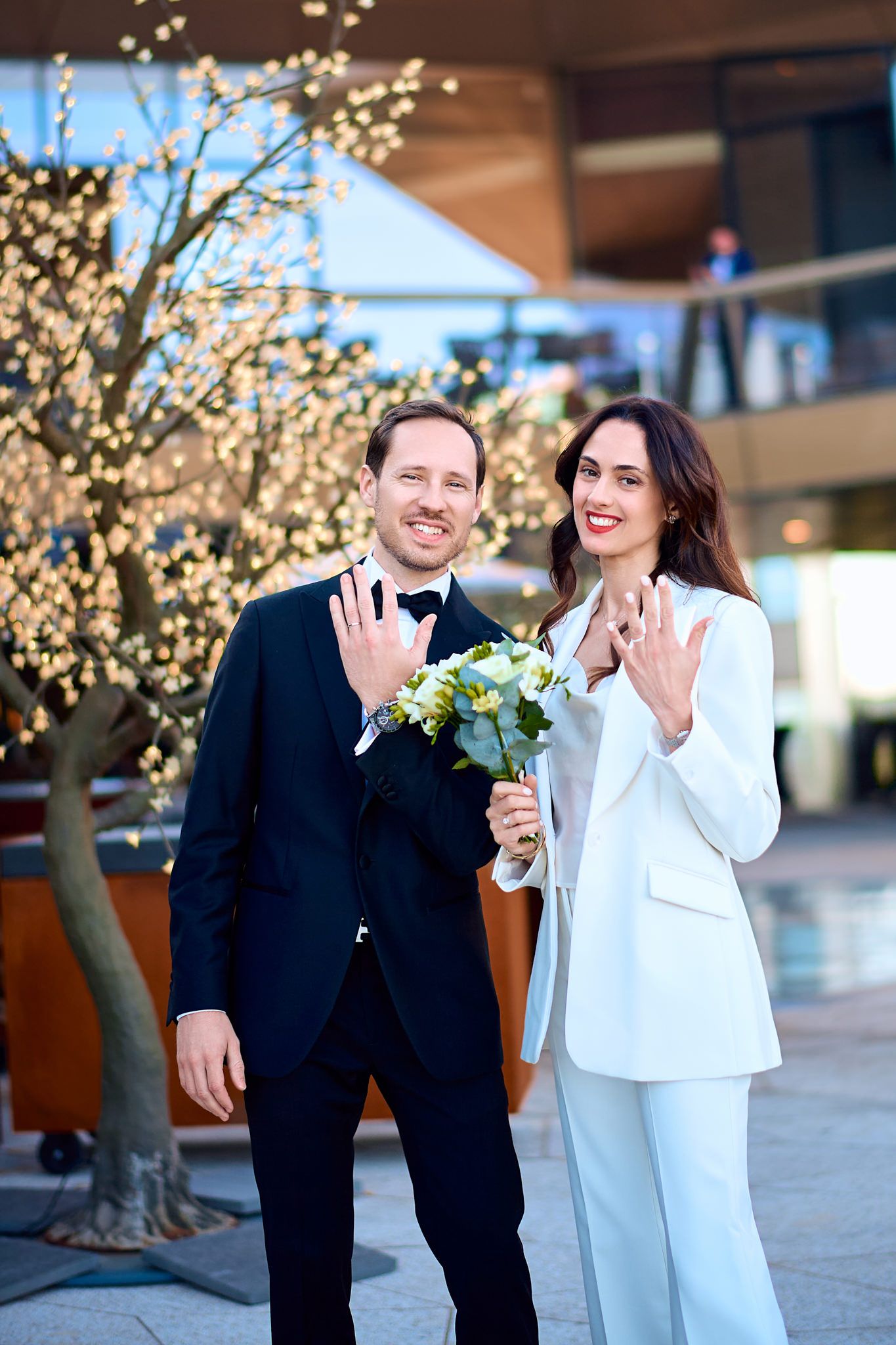 Couple in formal attire shows off rings near illuminated trees.