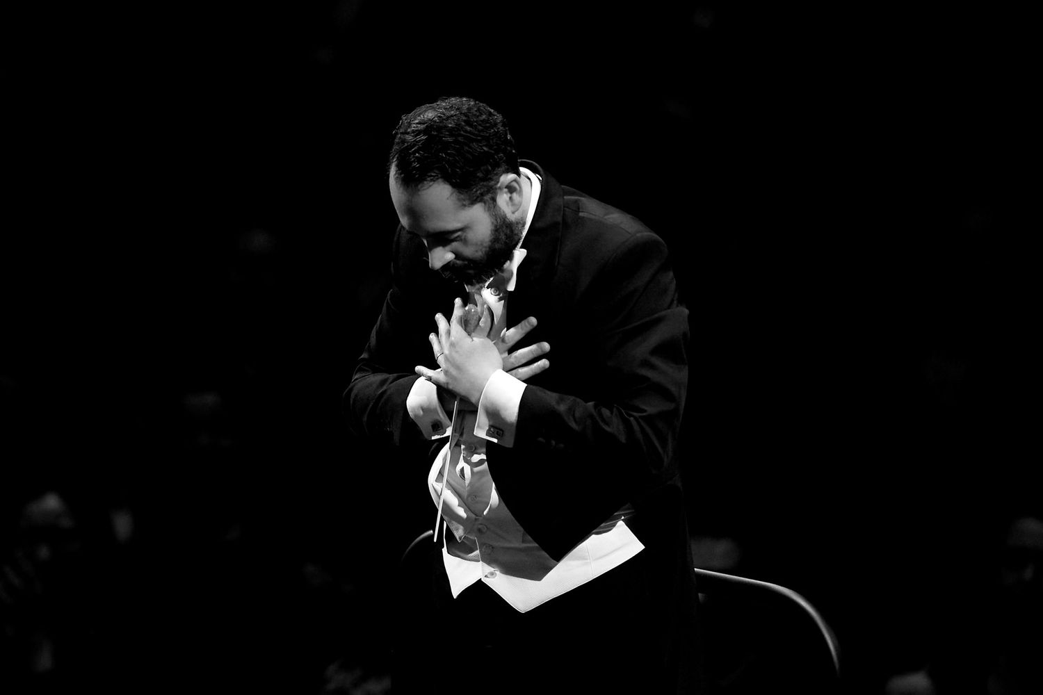 Conductor in formal tuxedo leads orchestra with dramatic gestures against dark background.