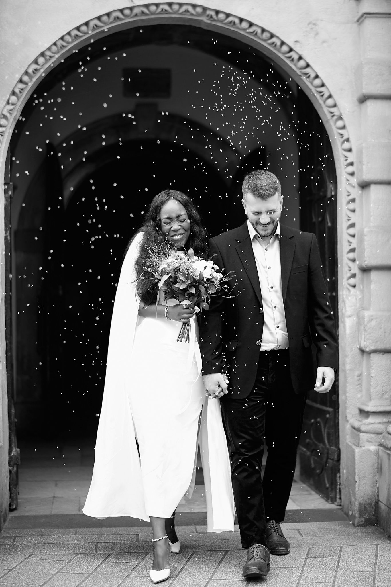Wedding party exits through stone archway while guests shower them with confetti.