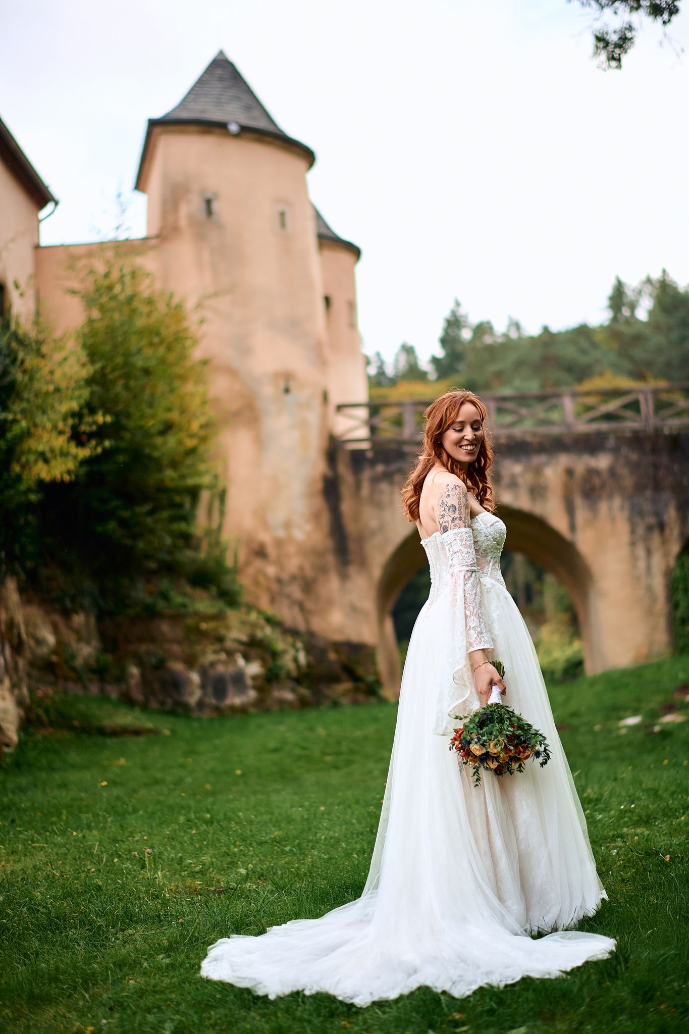 Bride in flowing white lace gown poses near historic castle grounds.