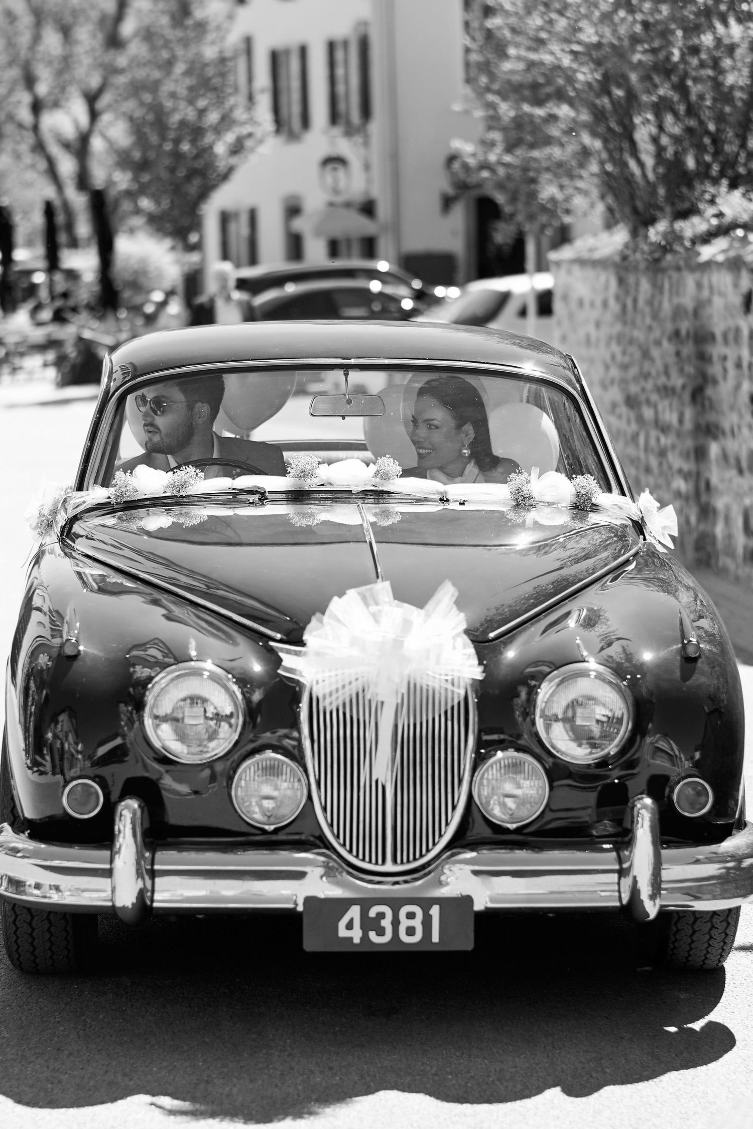 Bride and groom riding in a decorated vintage classic car with white bow, black and white wedding photo.