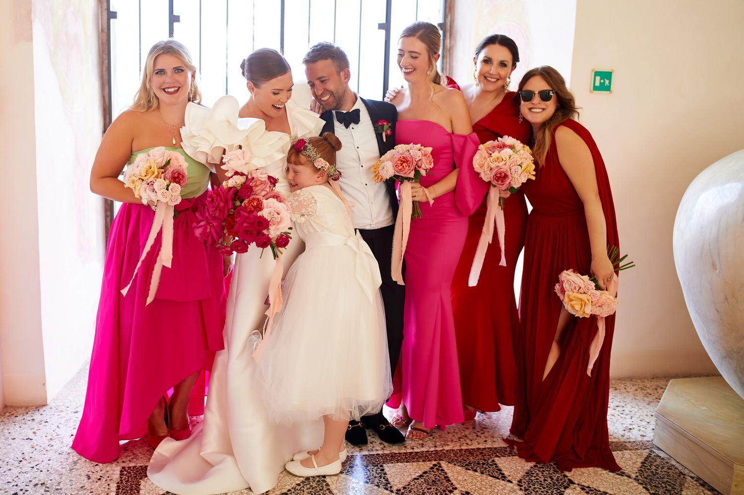 Wedding party in pink and burgundy dresses poses together indoors.