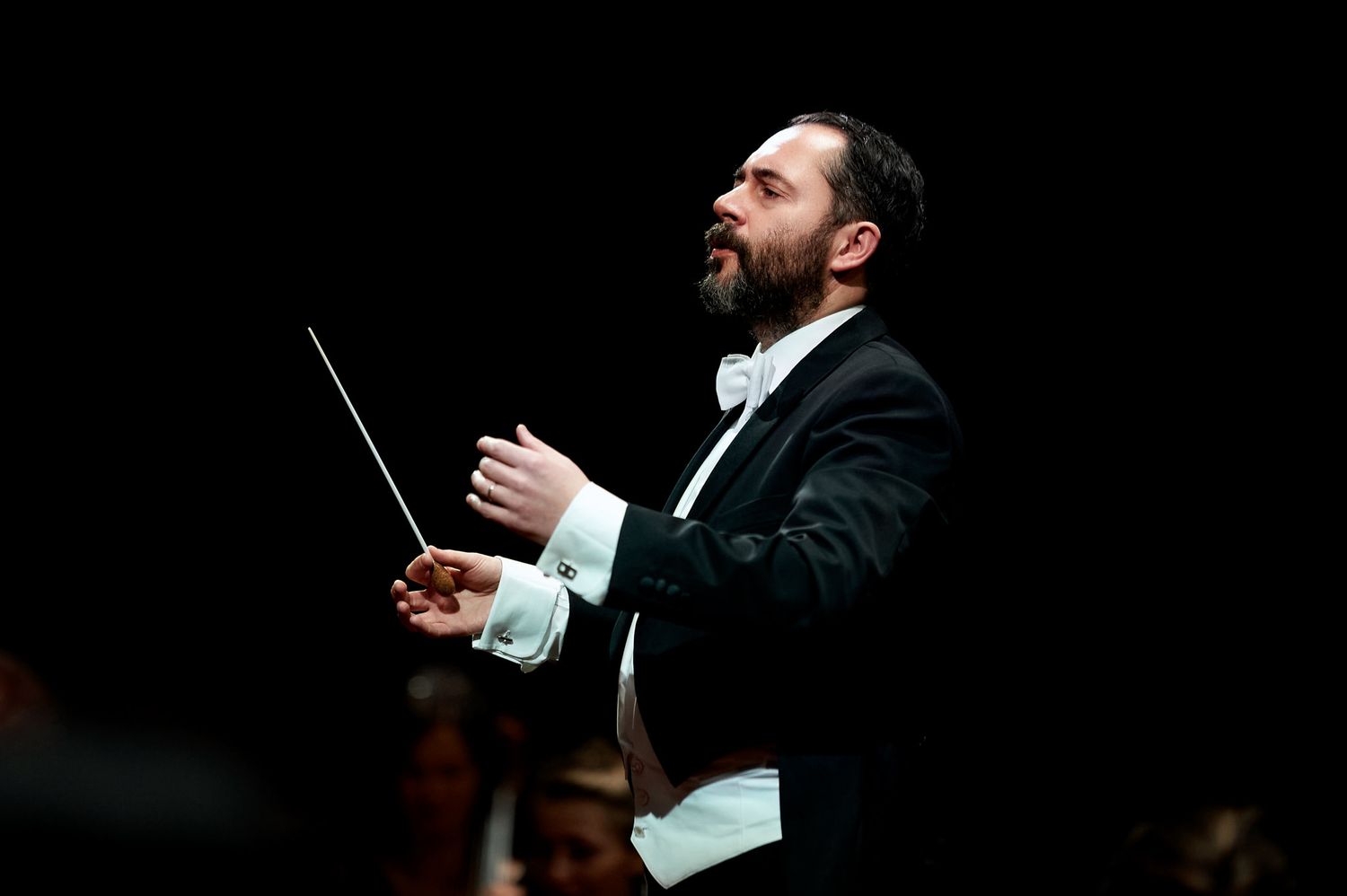 Orchestra conductor leads performance with dramatic gestures against dark background.