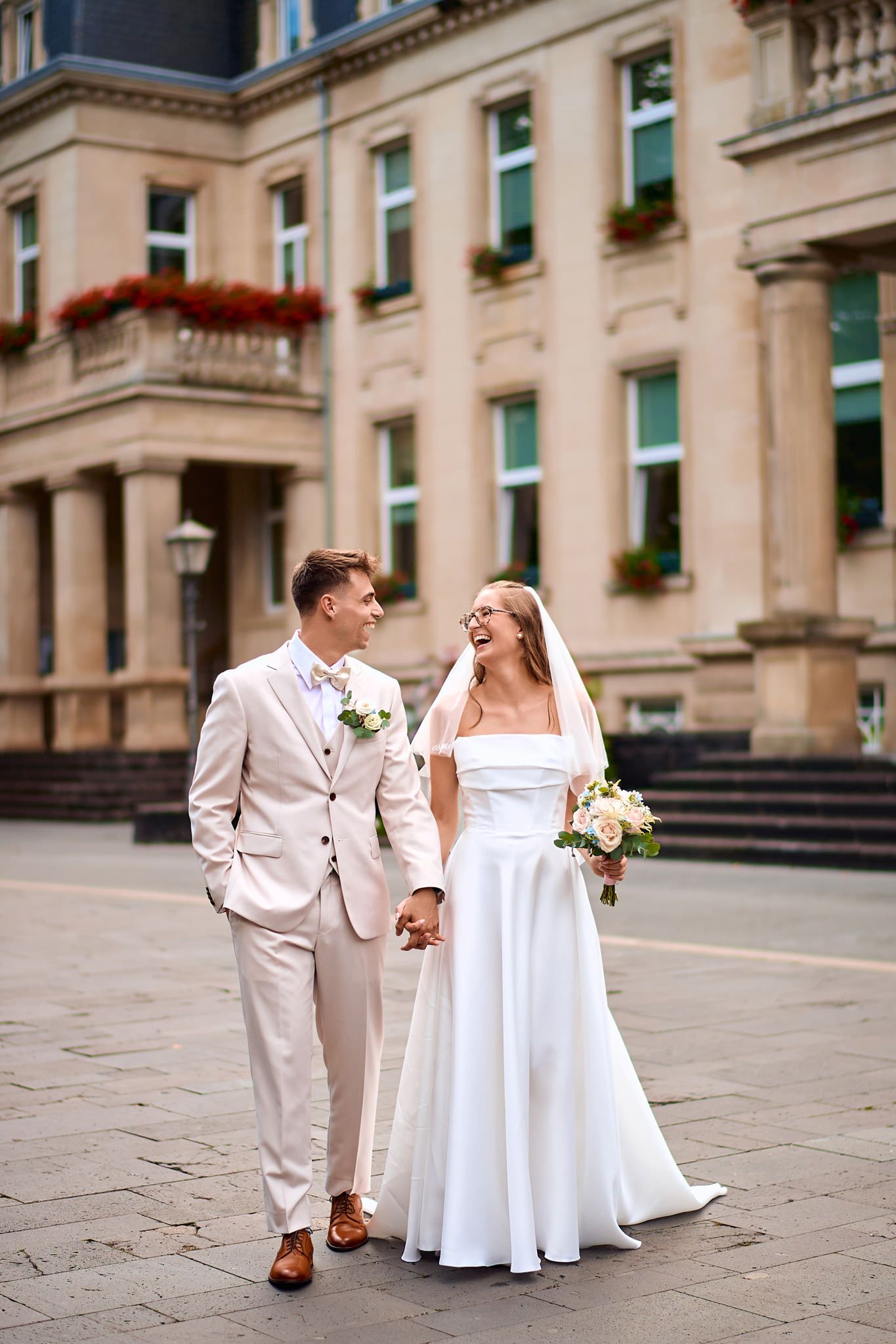 Wedding couple shares candid moments on stone plaza in front of elegant building.
