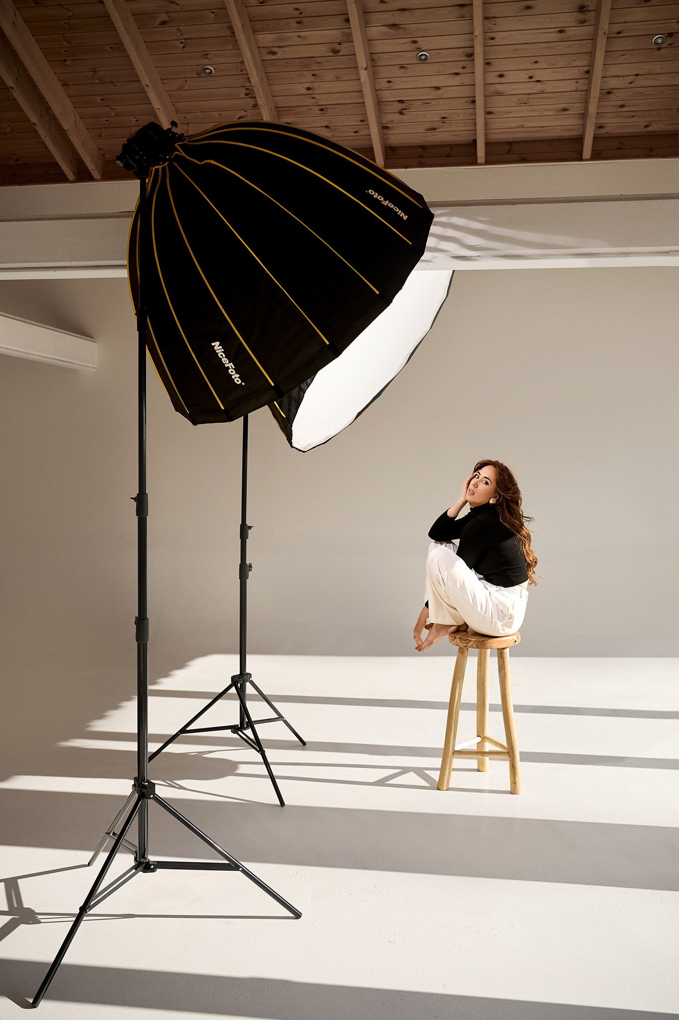 Professional photography studio setup with black umbrella lights and wooden stool in bright white room.