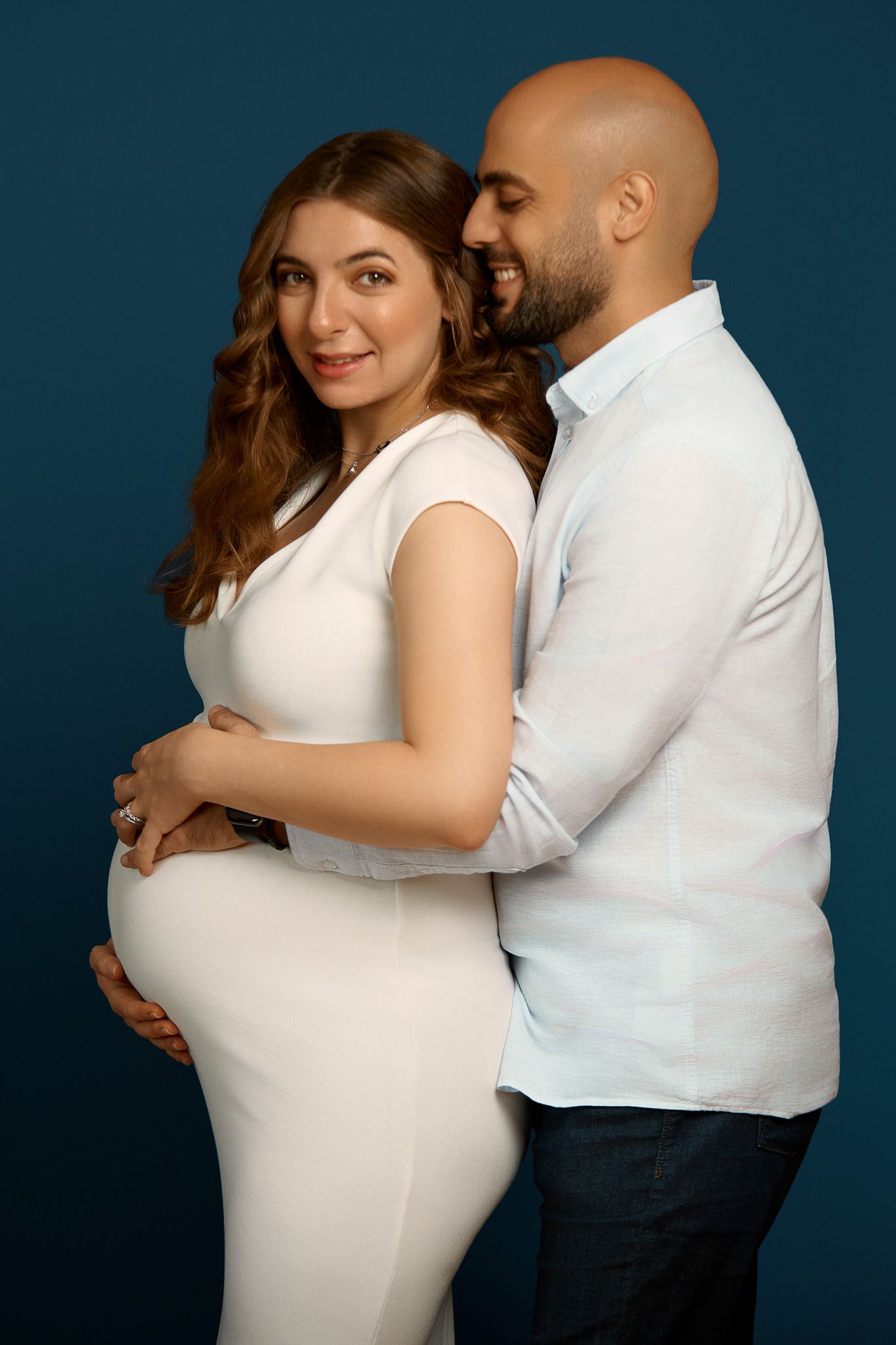 Romantic maternity portrait with couple embracing against deep blue studio background.