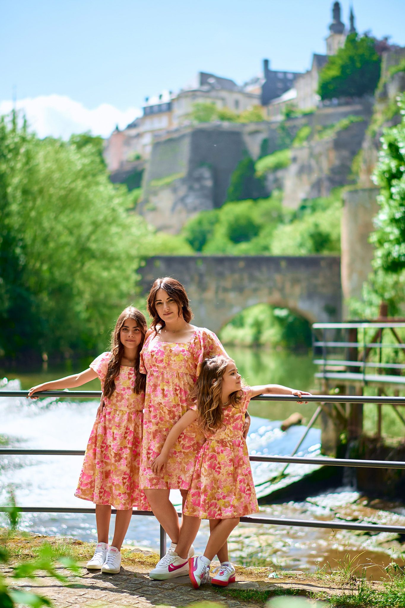 Children in pink floral dresses posing by a riverside railing with historic buildings in background.
