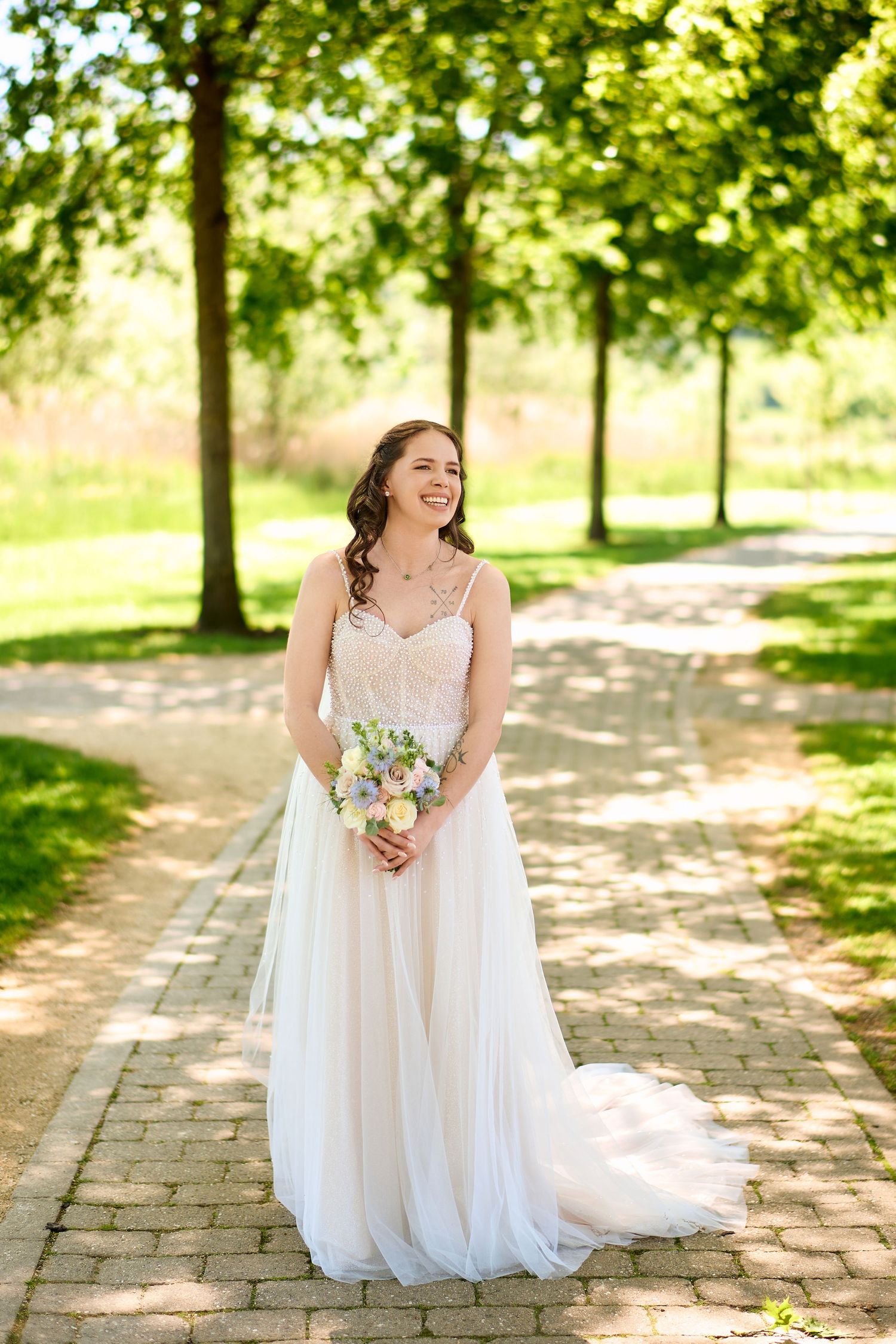 Smiling bride in white lace gown holding pastel bouquet on brick path surrounded by lush green trees in sunny park.