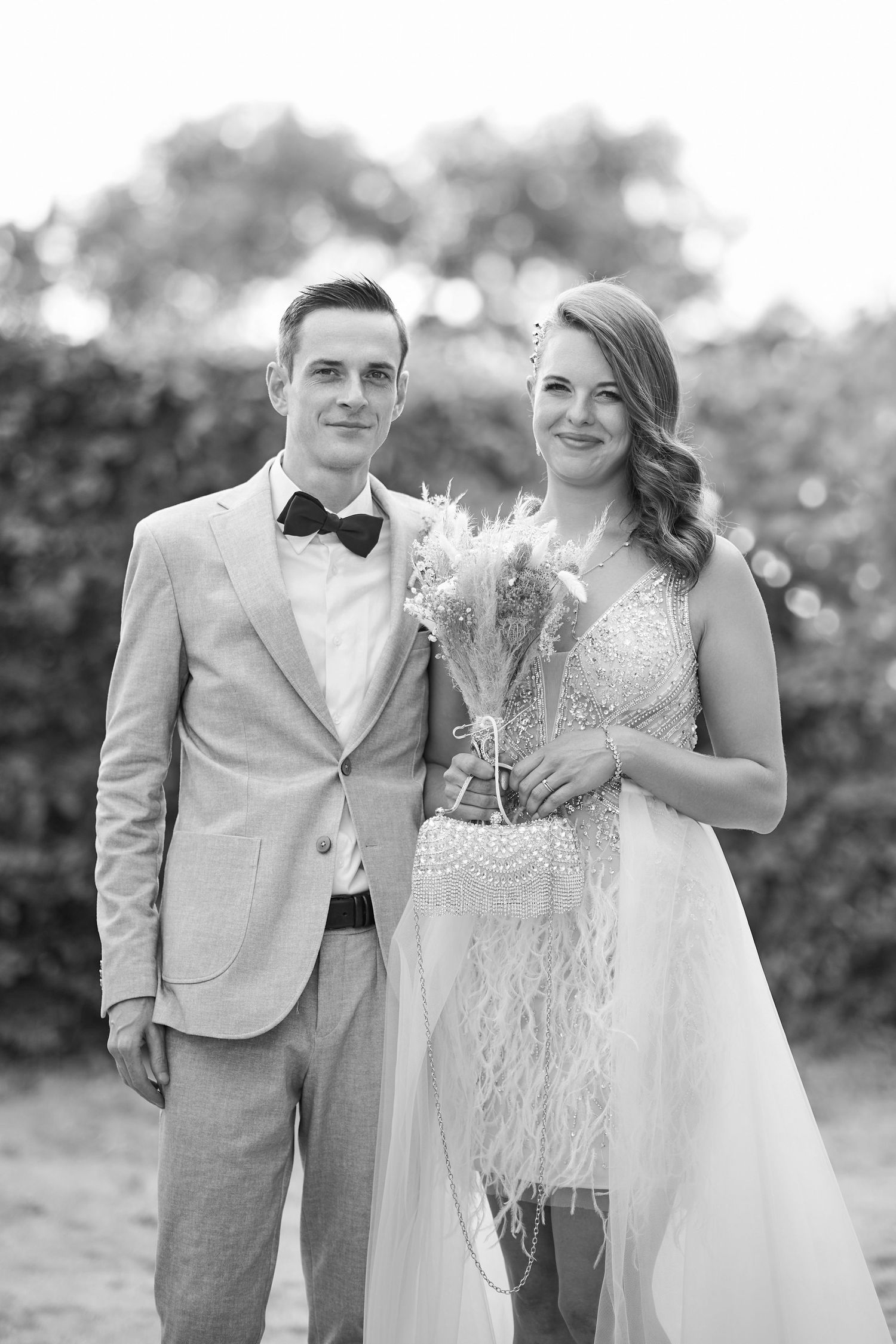 Black and white wedding portrait of smiling bride holding bouquet and groom in bow tie suit posing outdoors.