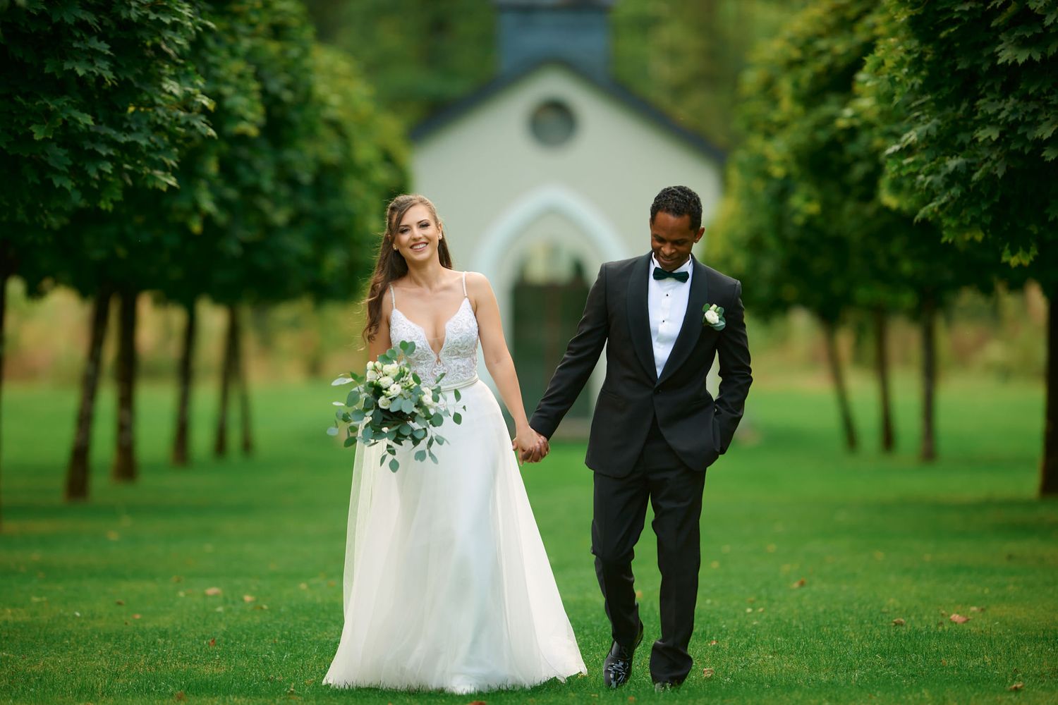 Wedding couple walks hand in hand on manicured lawn with chapel in background.