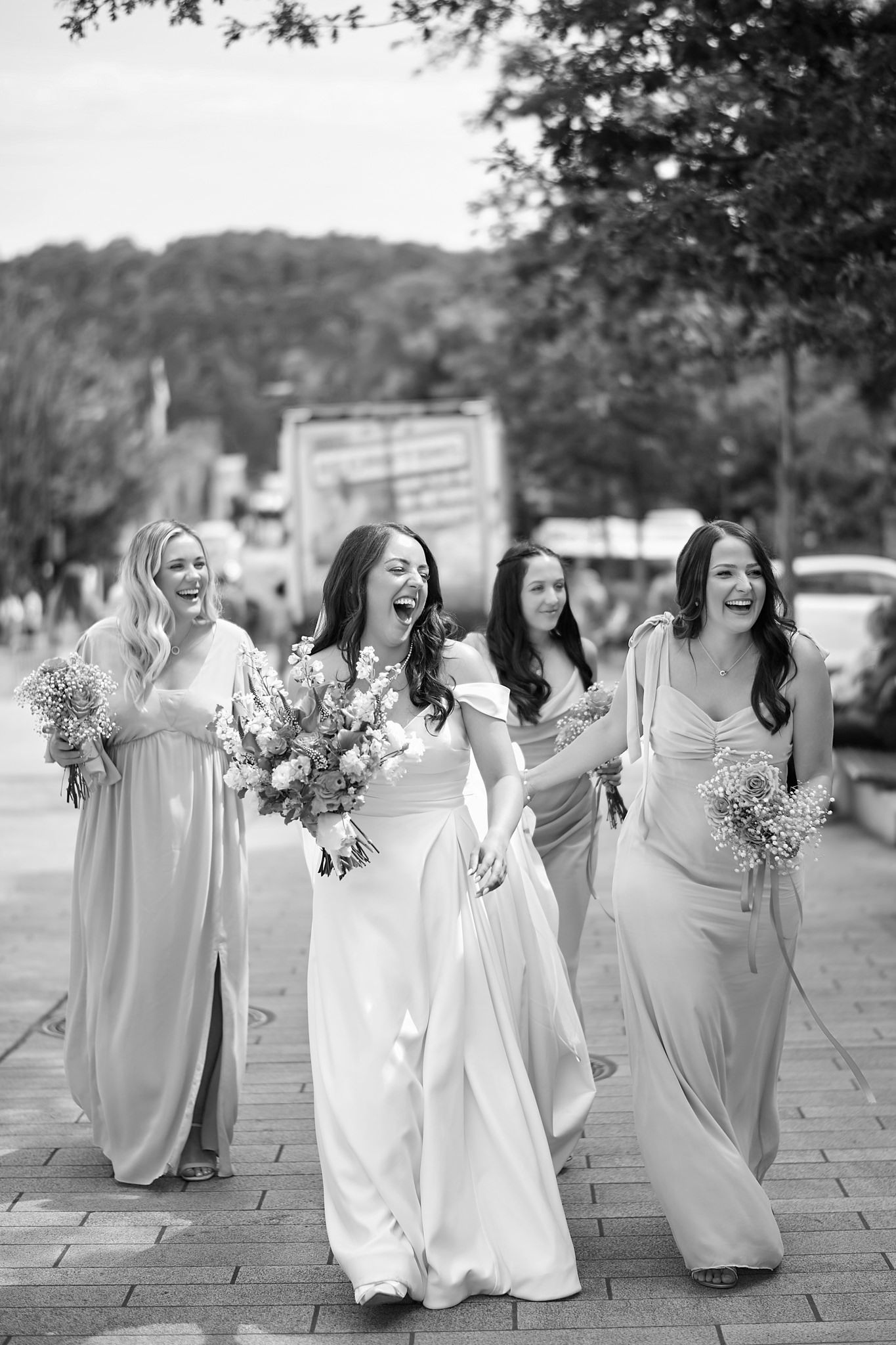 Bridesmaids in flowing white dresses walk together holding bouquets outdoors.