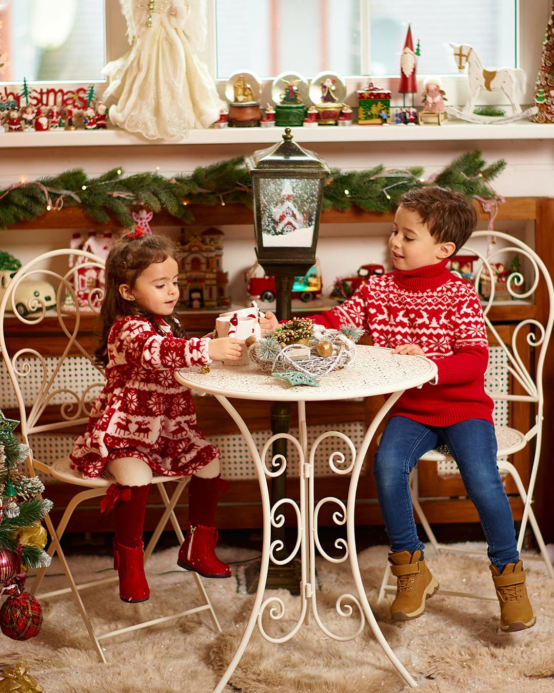 Two children in matching red Christmas sweaters sit at a white ornate table surrounded by festive holiday decorations.