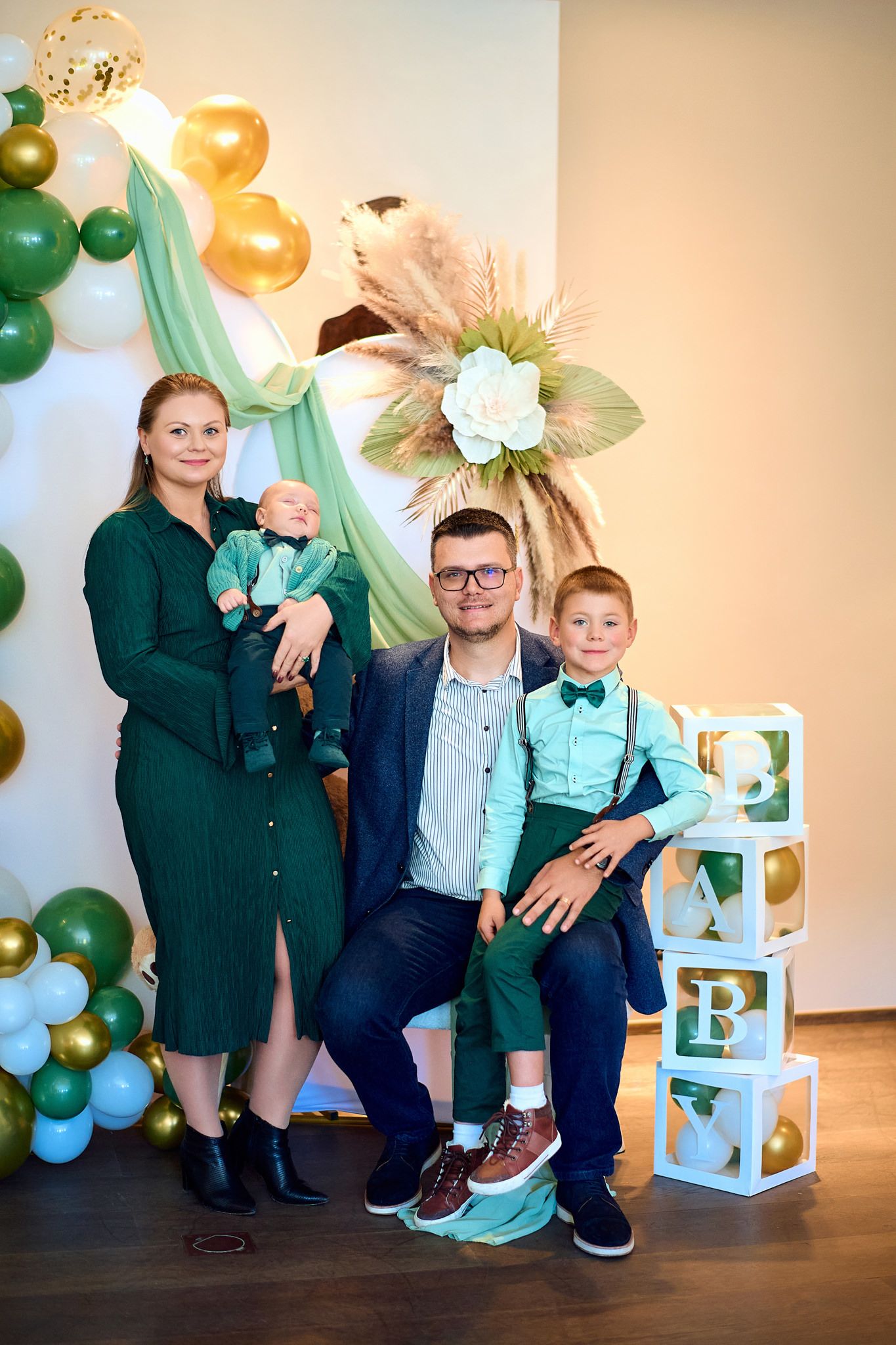 Mother in green dress holds sleeping baby against backdrop of festive balloon decorations.