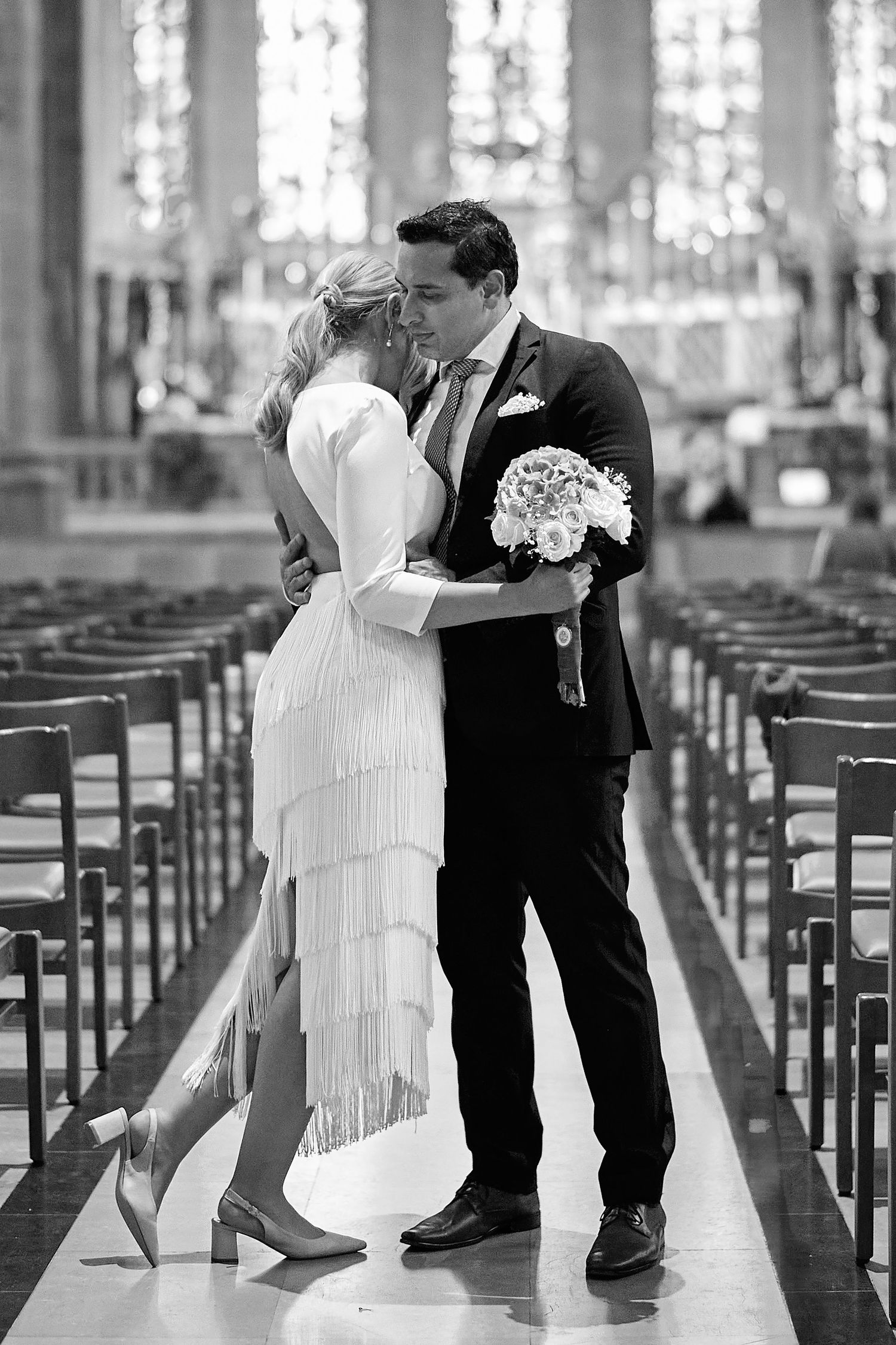 A romantic wedding couple shares a kiss in an elegant church aisle surrounded by wooden pews.