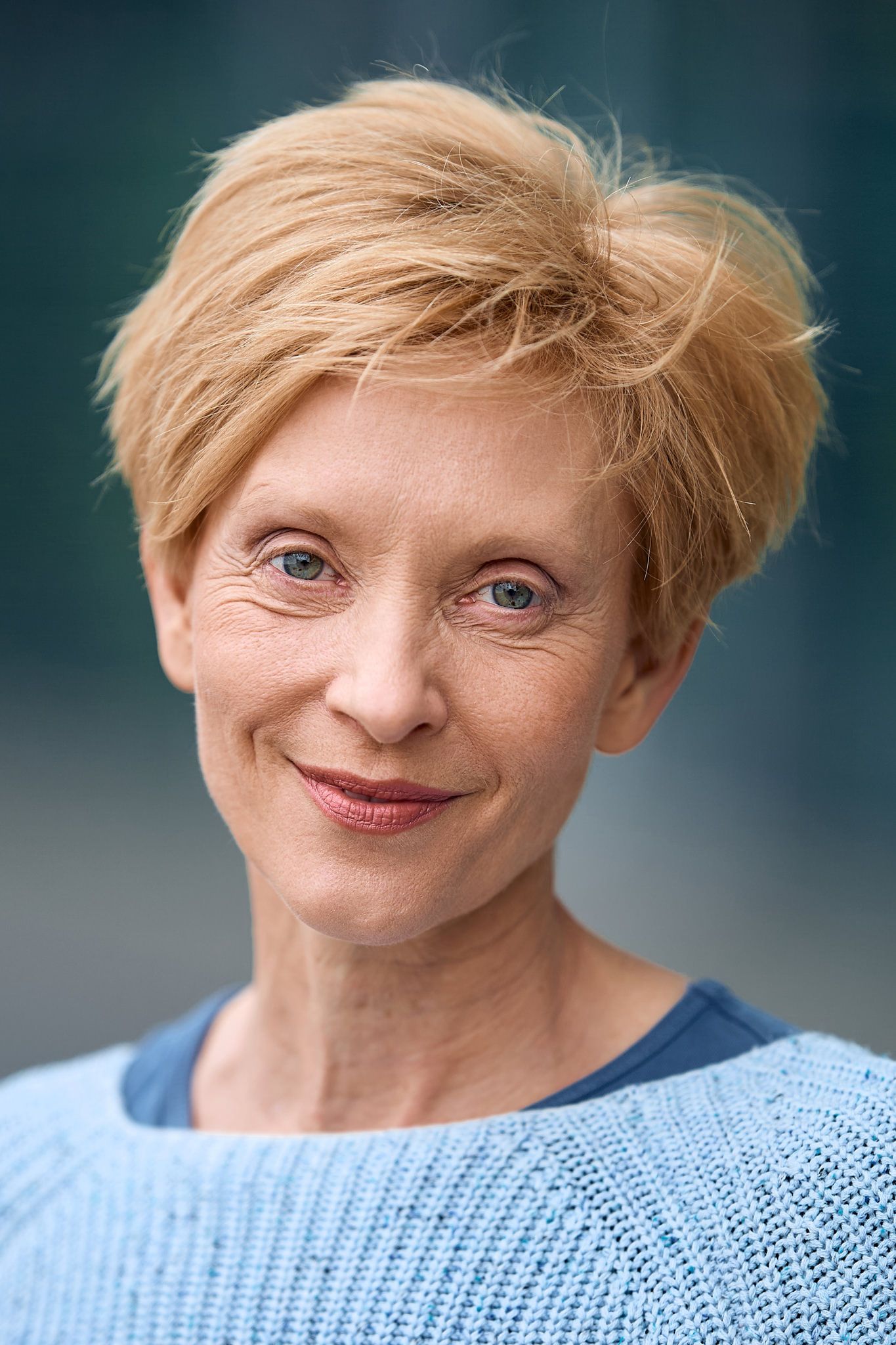 Woman in light blue sweater with short blonde hair smiling against dark background.
