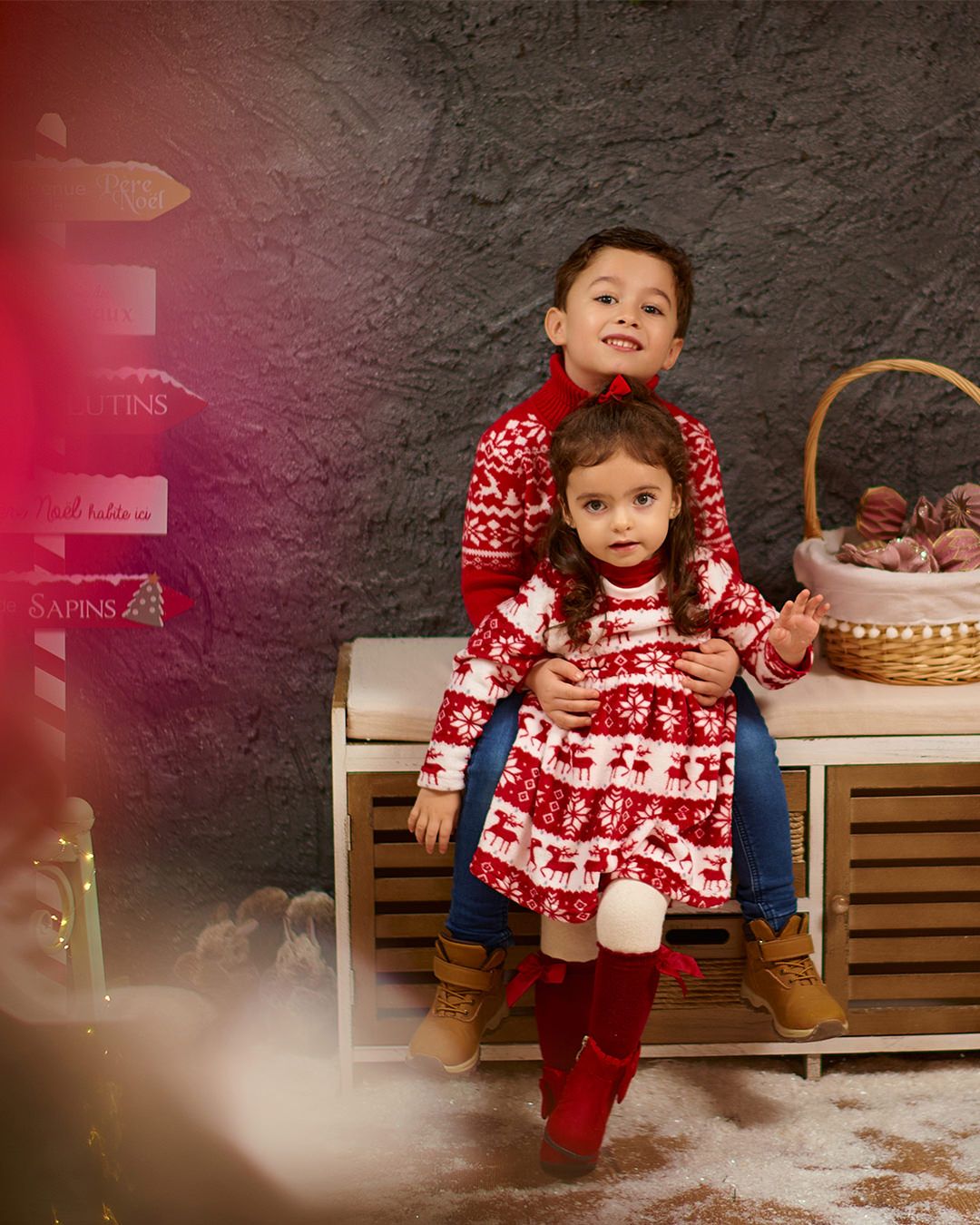 Two children in matching red and white Nordic Christmas sweaters pose together on a wooden bench with holiday decorations.