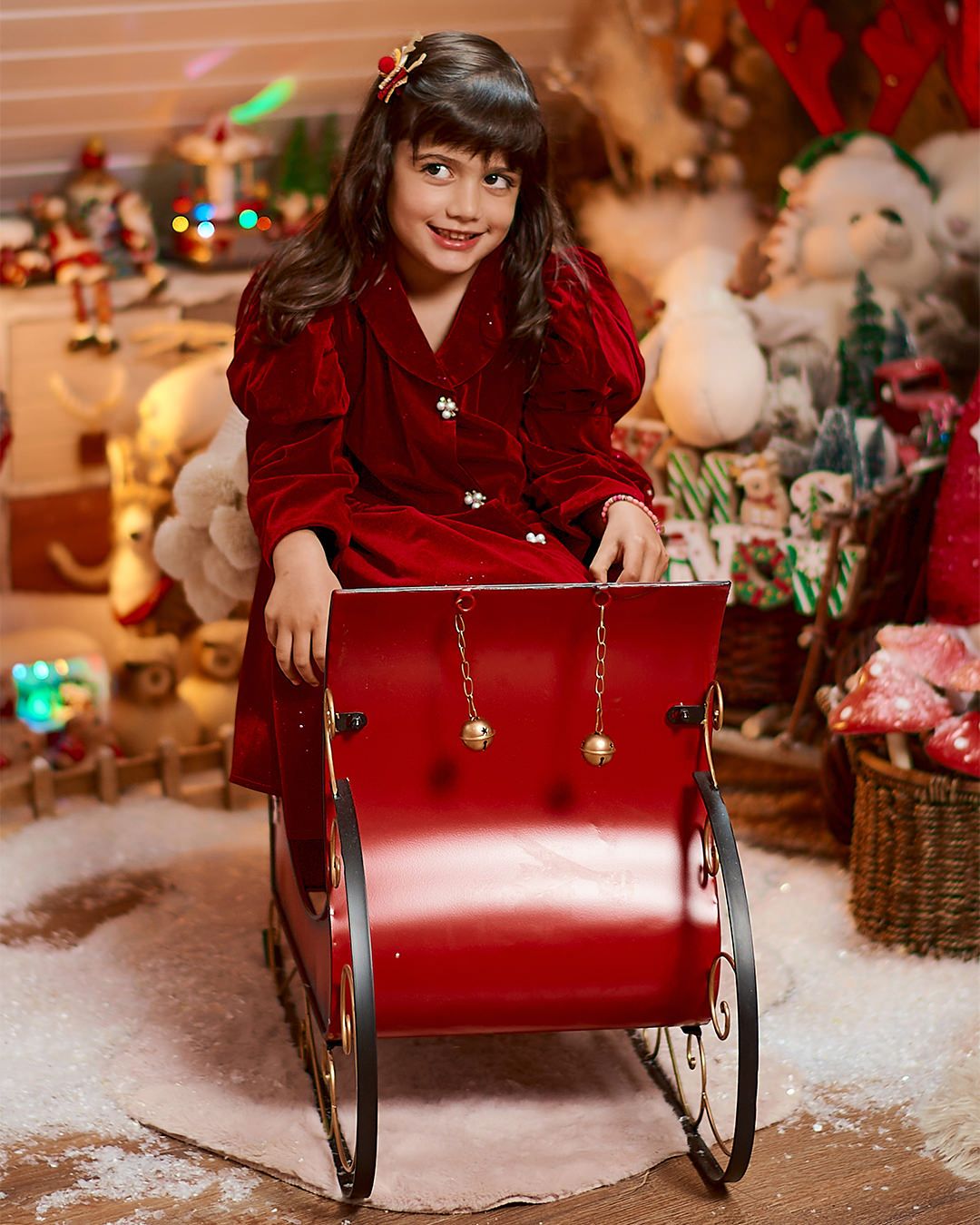 Young girl in red velvet dress sitting in a red Christmas sleigh prop surrounded by festive holiday decorations.