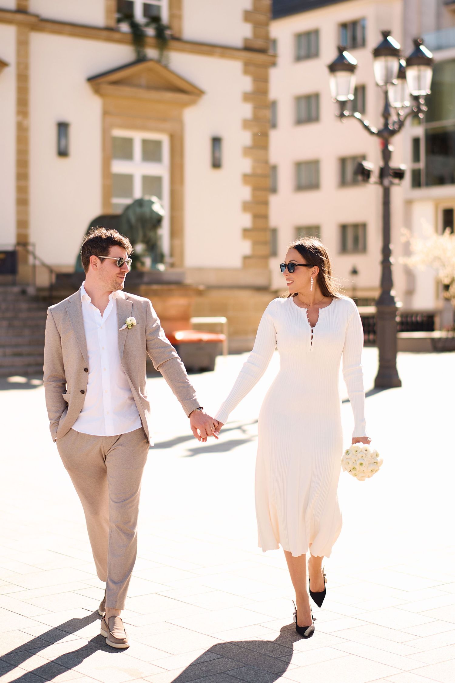 Elegant couple in wedding attire holding hands while walking through a sunny European city square.
