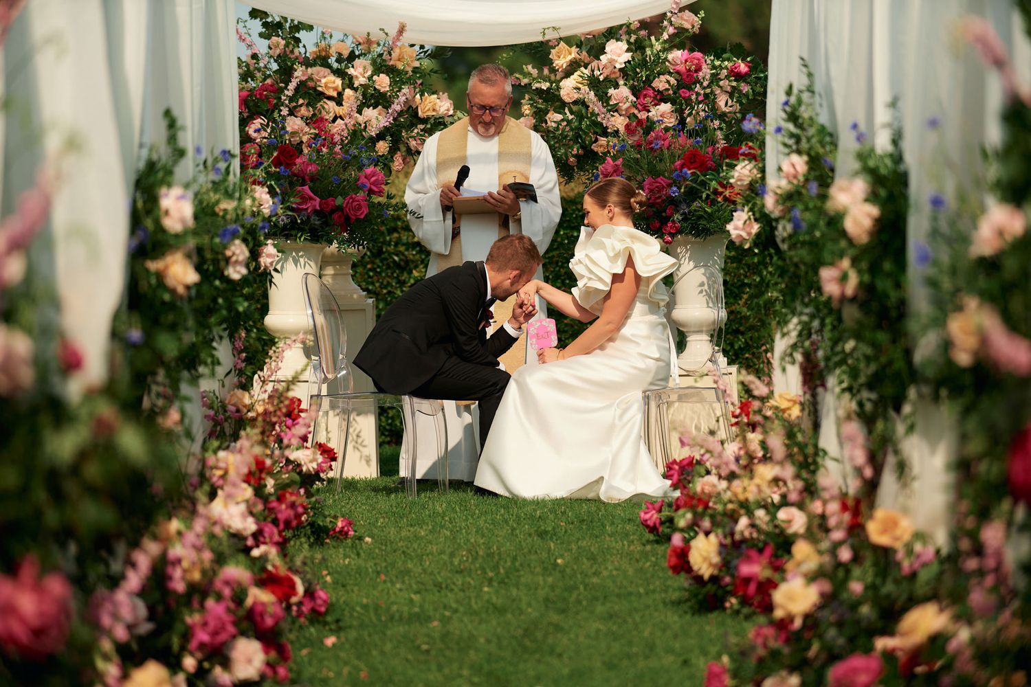 Elegant wedding ceremony setup with white drapes and pink floral arrangements in a garden setting.