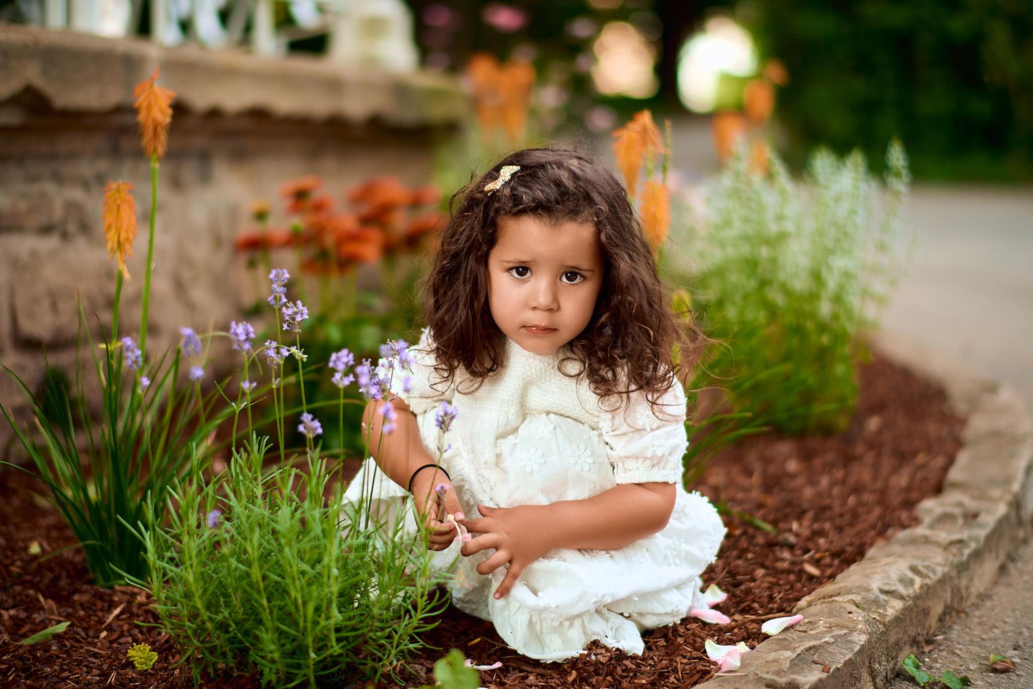 Child in white dress sitting among garden flowers and greenery.