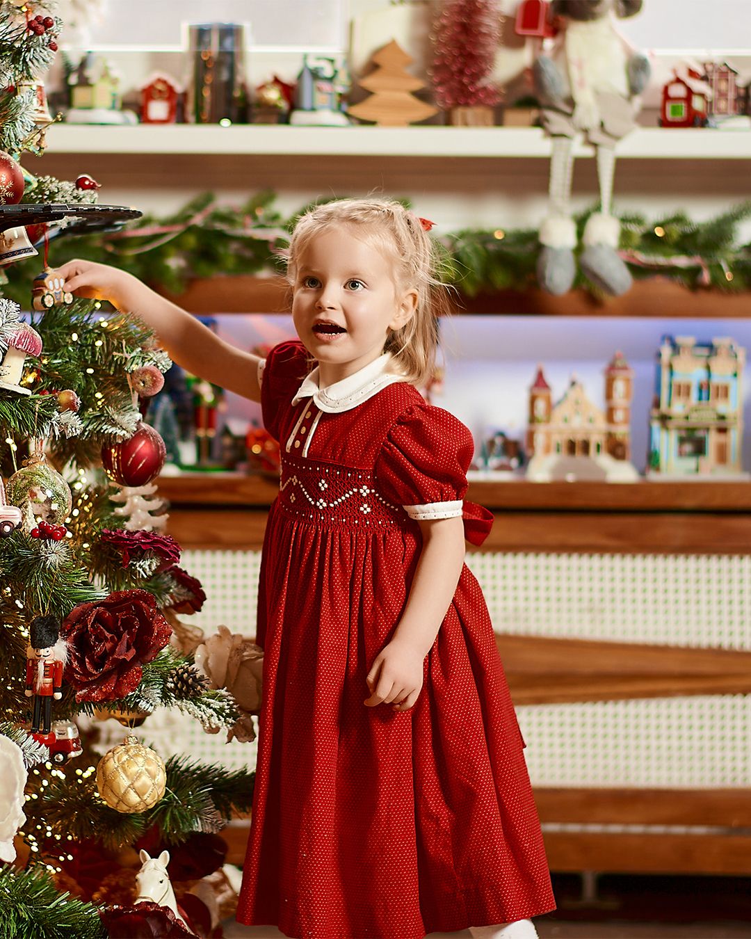 Toddler girl in red smocked dress reaching to decorate a Christmas tree in a festive holiday room.