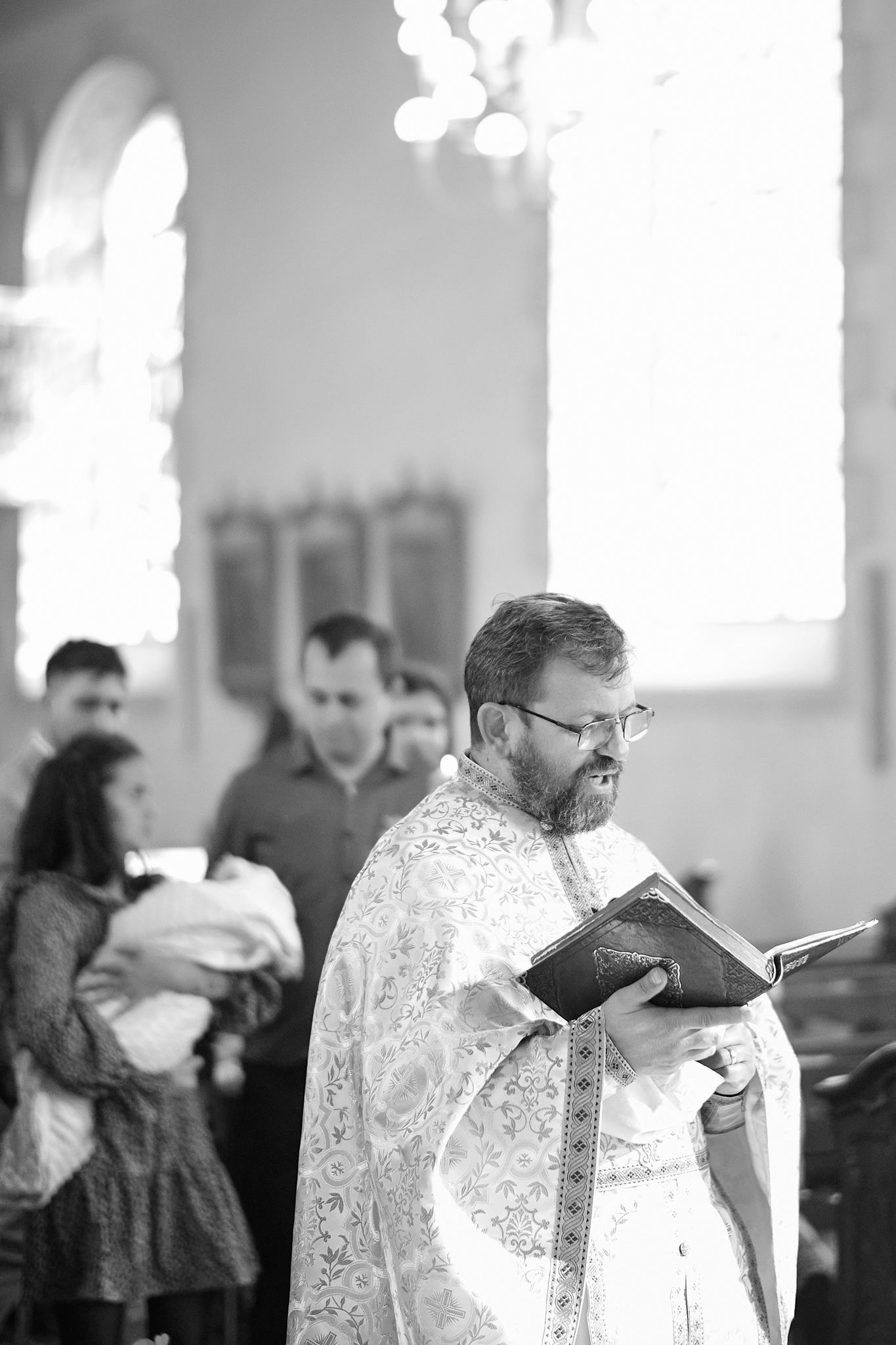 Orthodox priest in white embroidered vestments reads from a religious text during a church service.