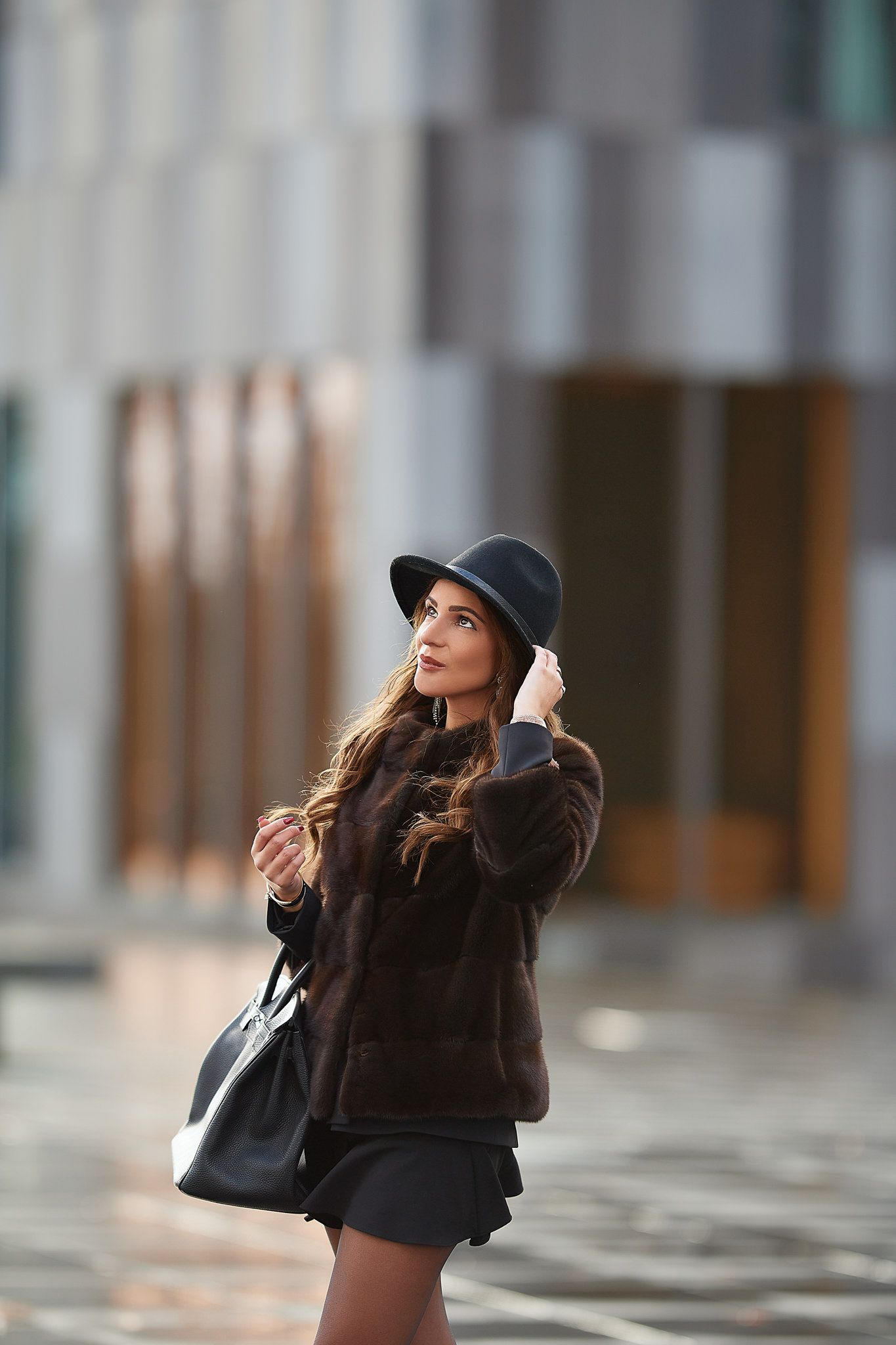 Stylish figure in black hat and coat poses in modern architectural setting with dramatic lighting.