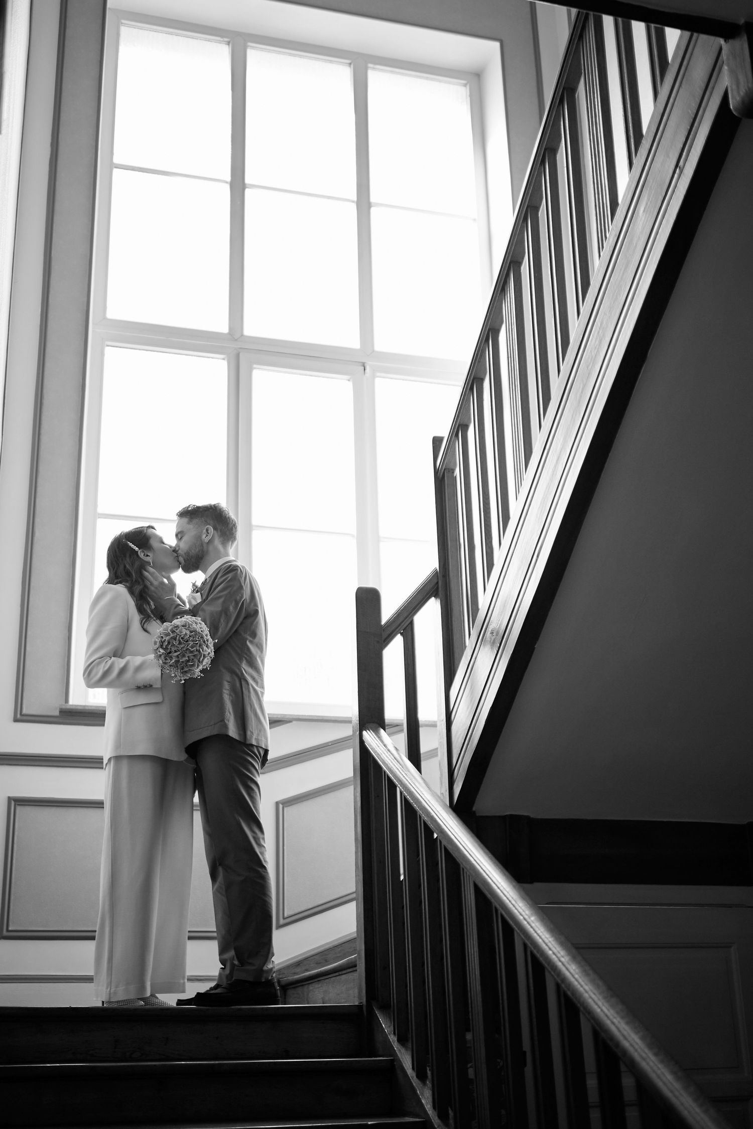 Black and white photo of a couple sharing a romantic kiss on a staircase in front of a large window.