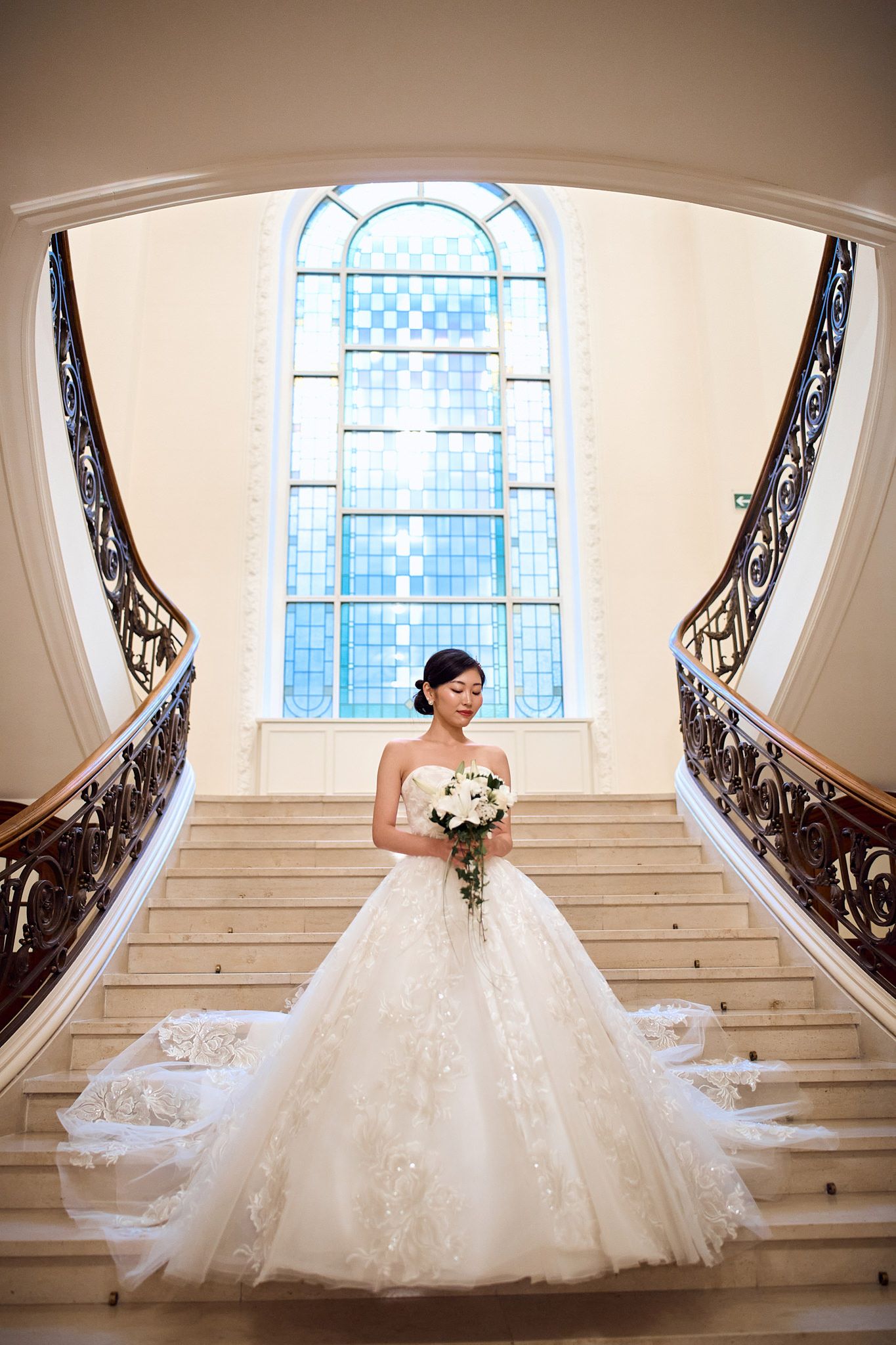 Dramatic bridal portrait on elegant curved staircase with stained glass window backdrop.