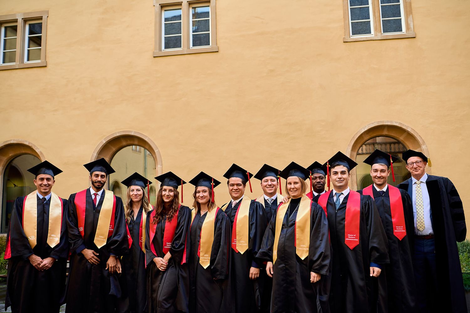 Large group of graduates in academic regalia lined up against a beige building wall.