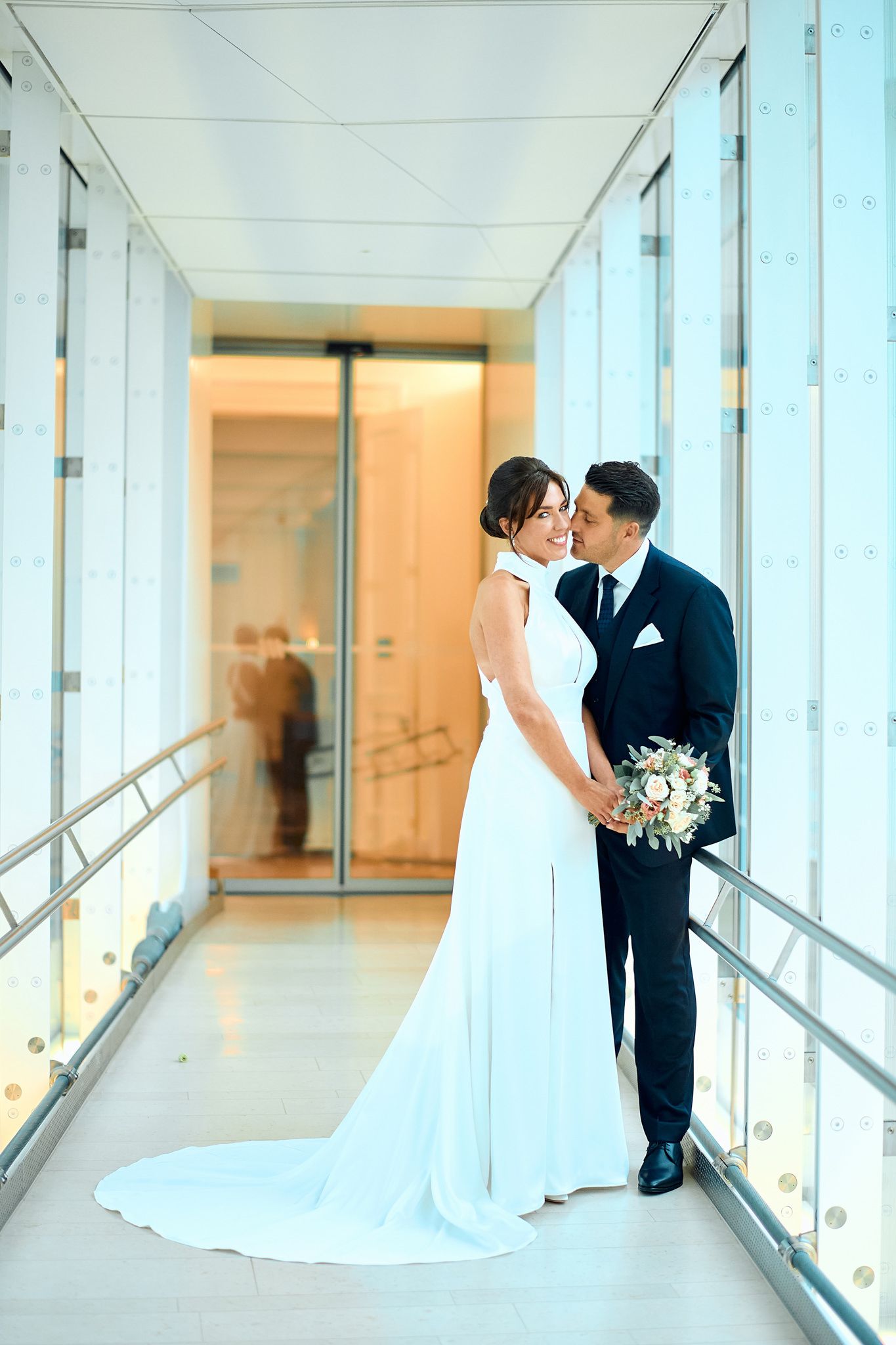 Wedding couple shares romantic moments in a bright modern hallway with floor to ceiling windows.