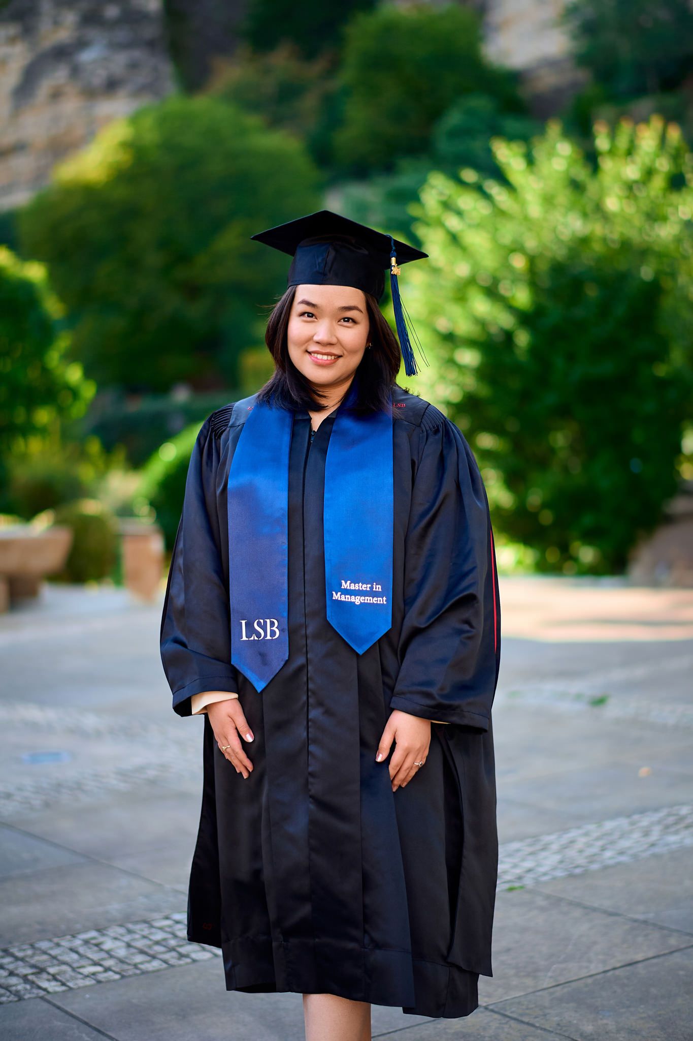 Graduate in cap and gown poses outdoors on campus with blue graduation stole.
