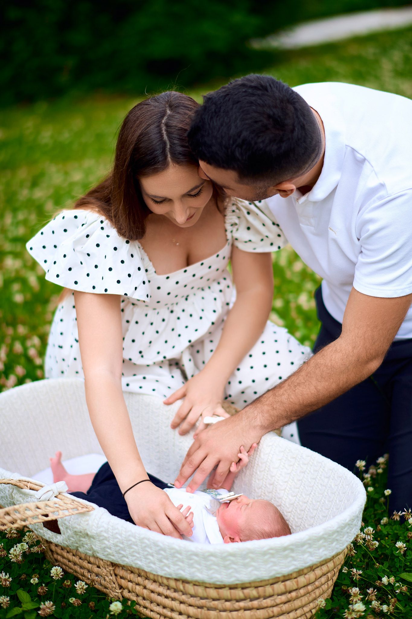Parents in white and polka dot outfits tend to baby in woven bassinet outdoors.