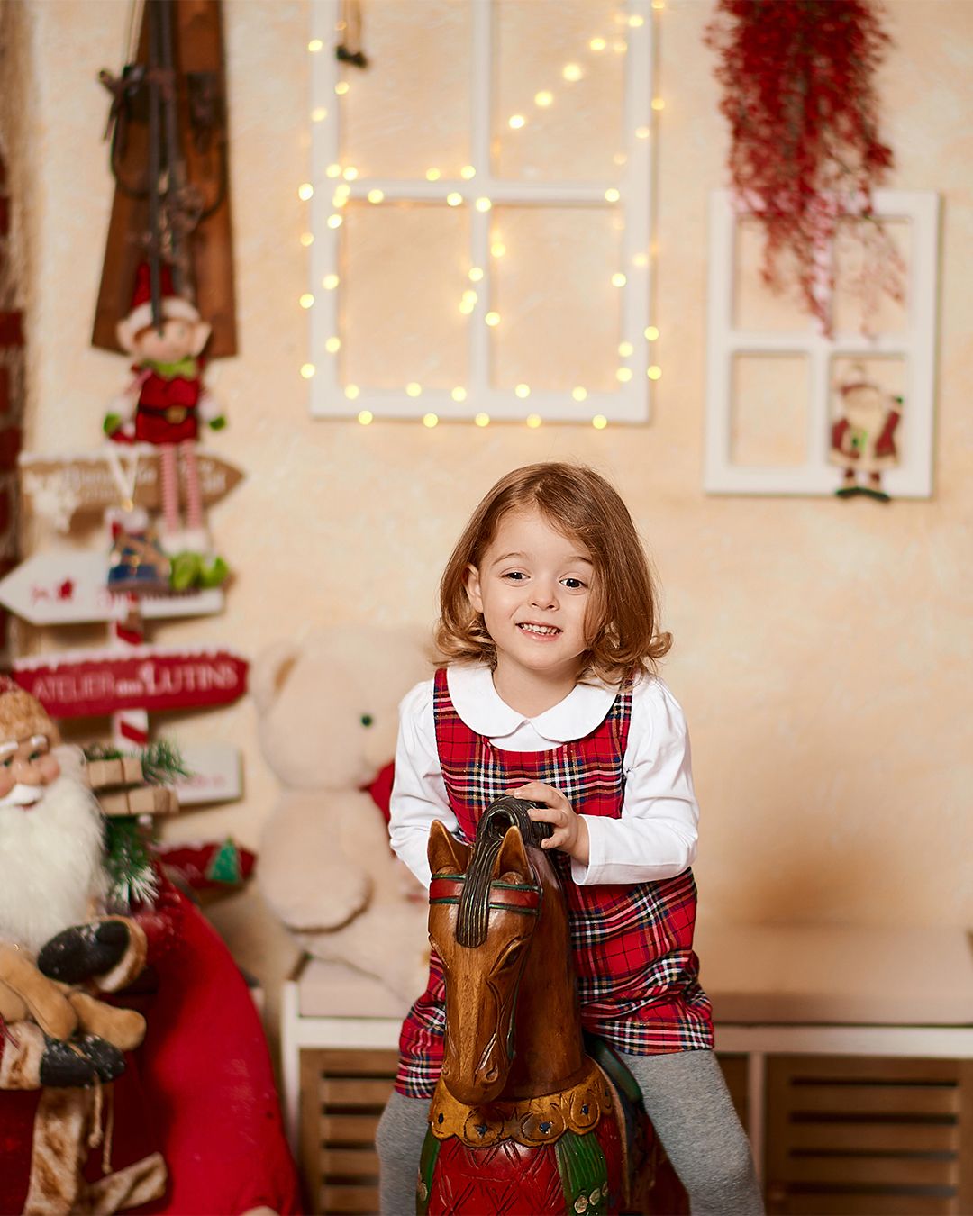 A smiling toddler girl in red plaid dress rides a wooden rocking horse in a festive Christmas-decorated room.