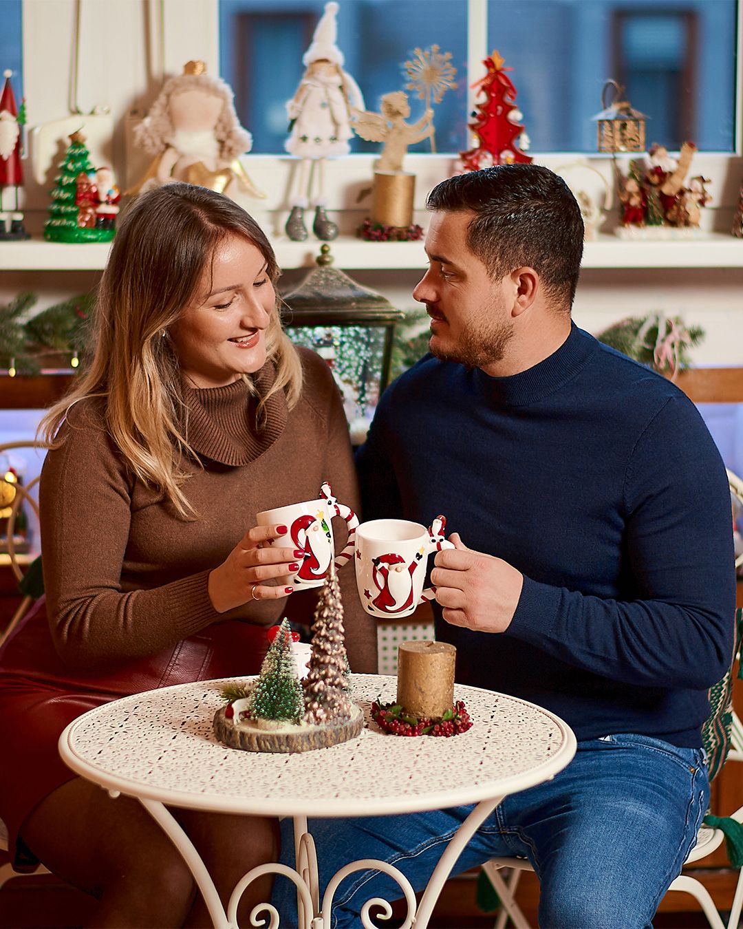 Couple smiling and clinking Christmas mugs at a festive table with holiday decorations in the background.