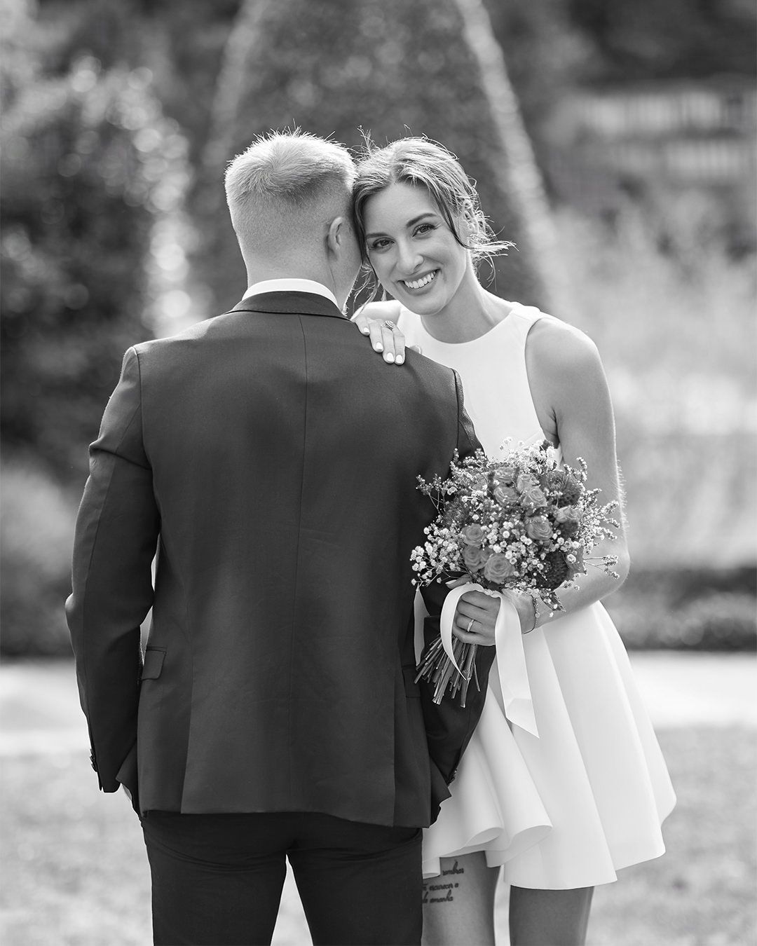 Black and white photo of a smiling bride in short white dress holding bouquet, facing groom in dark suit outdoors.