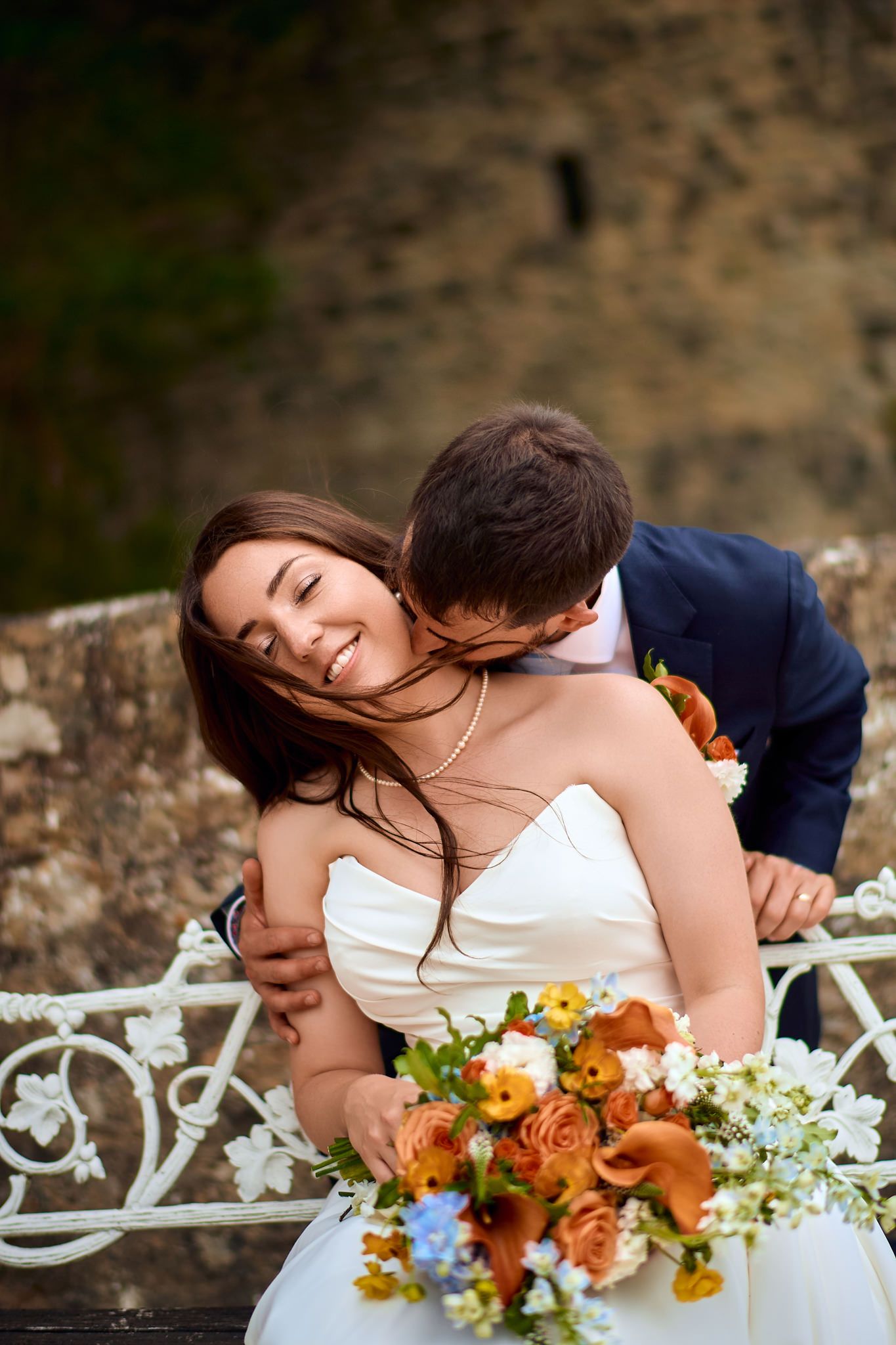 A romantic moment captured against a stone wall with orange and white floral bouquet accents.