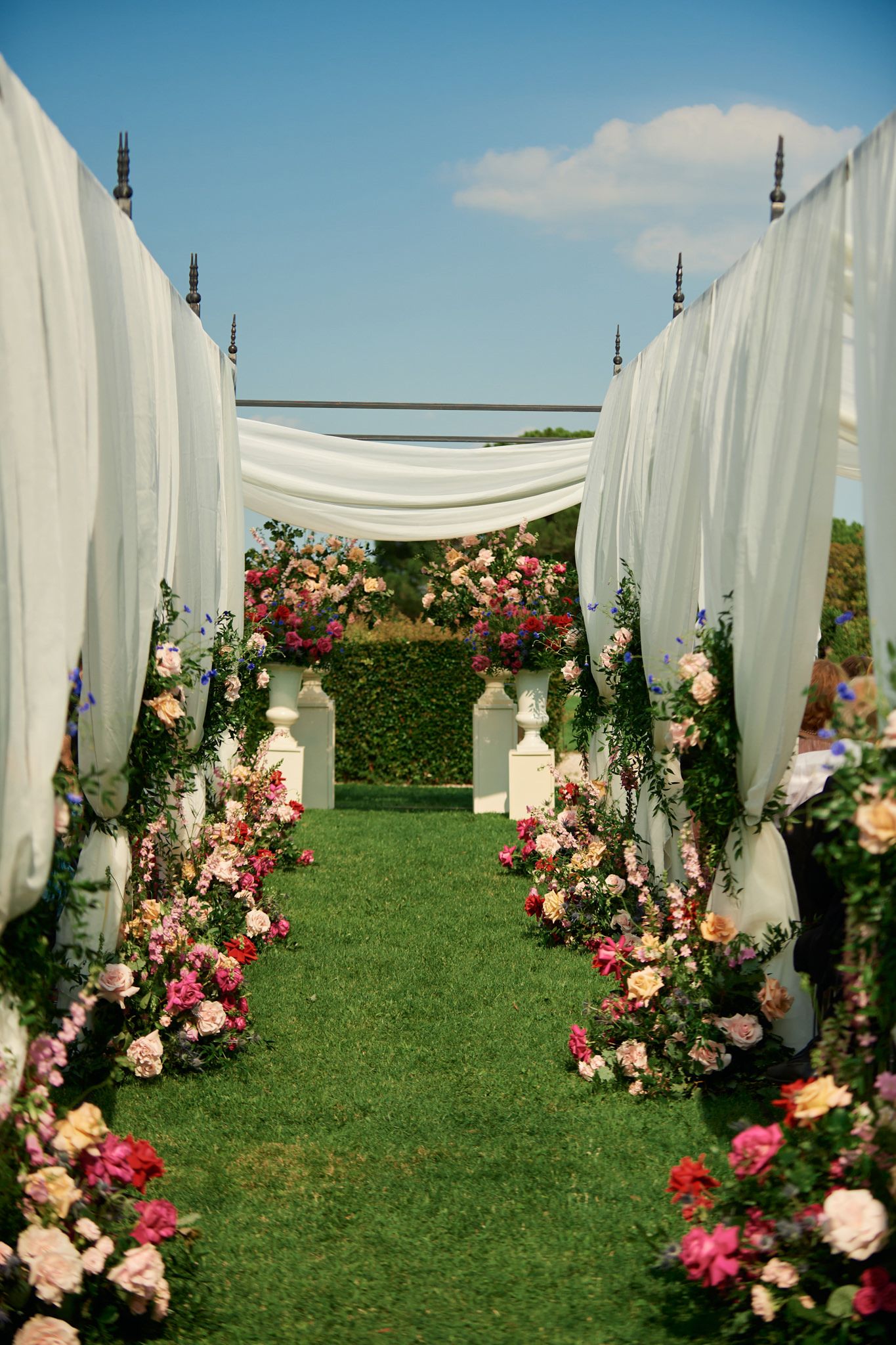 White fabric drapes and pink floral arrangements create an outdoor wedding aisle.
