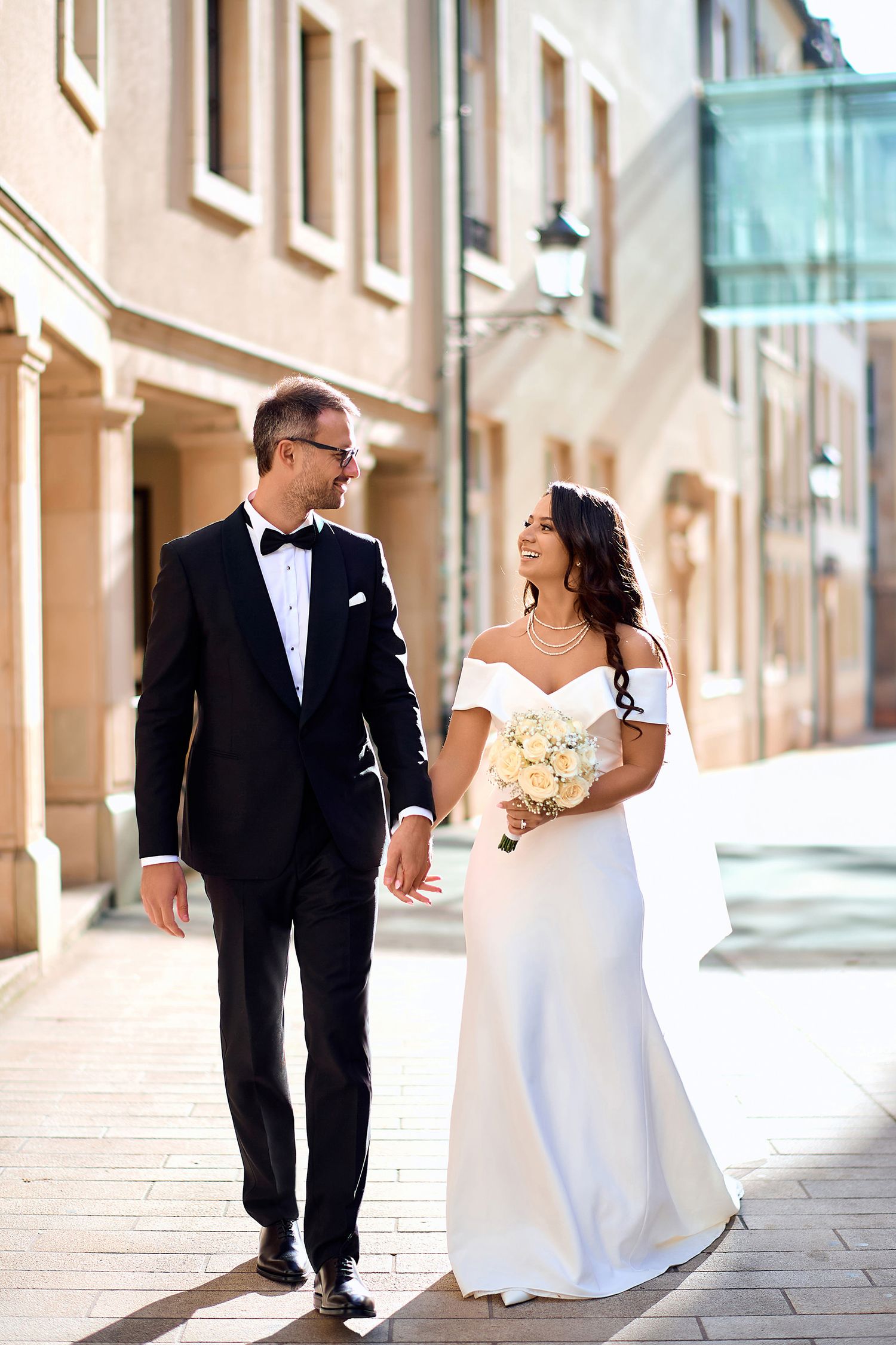 Elegant wedding couple walking on cobblestone street with bride holding white bouquet.