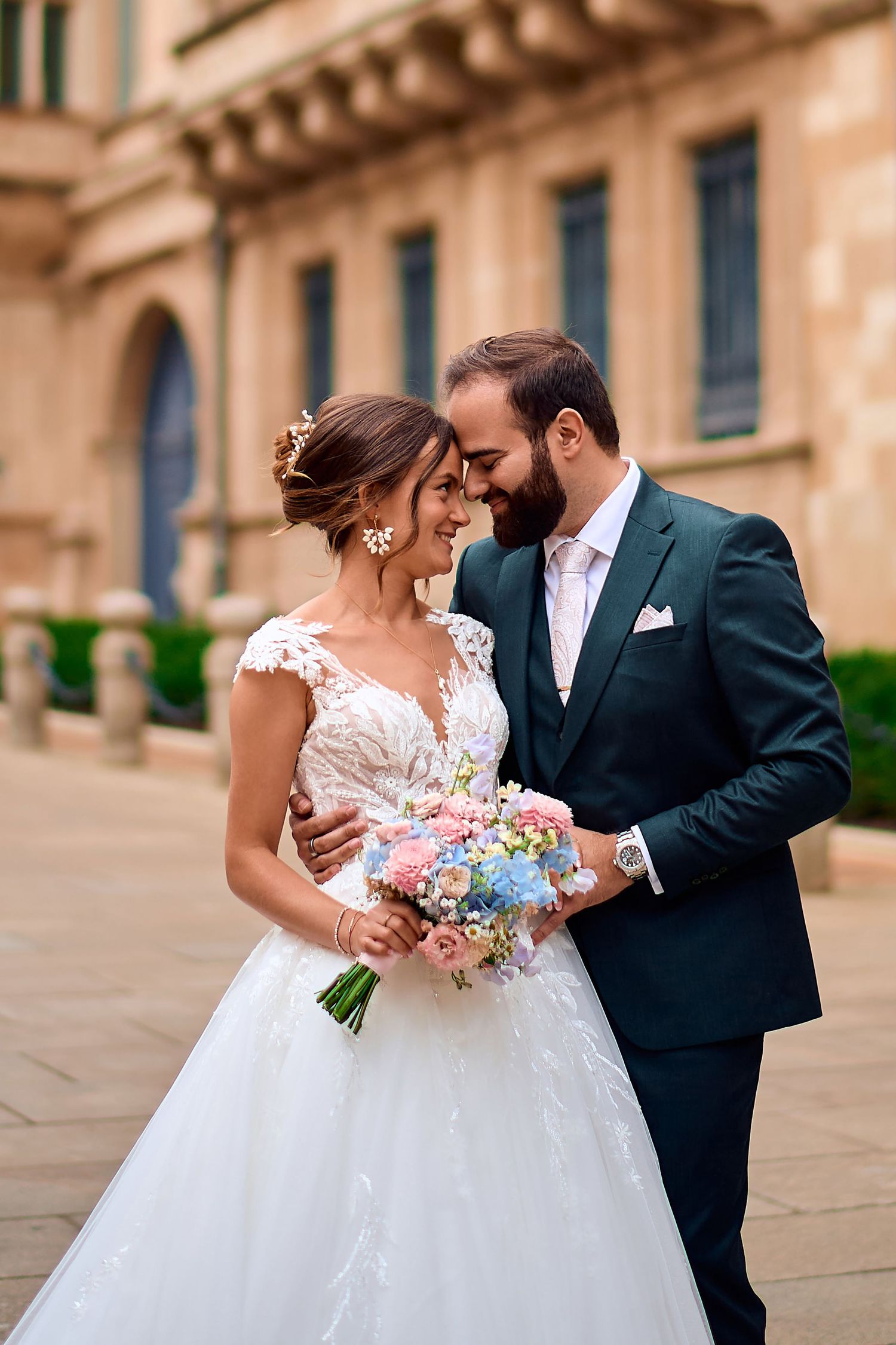 Romantic wedding couple embracing with bride holding pastel bouquet against stone building.