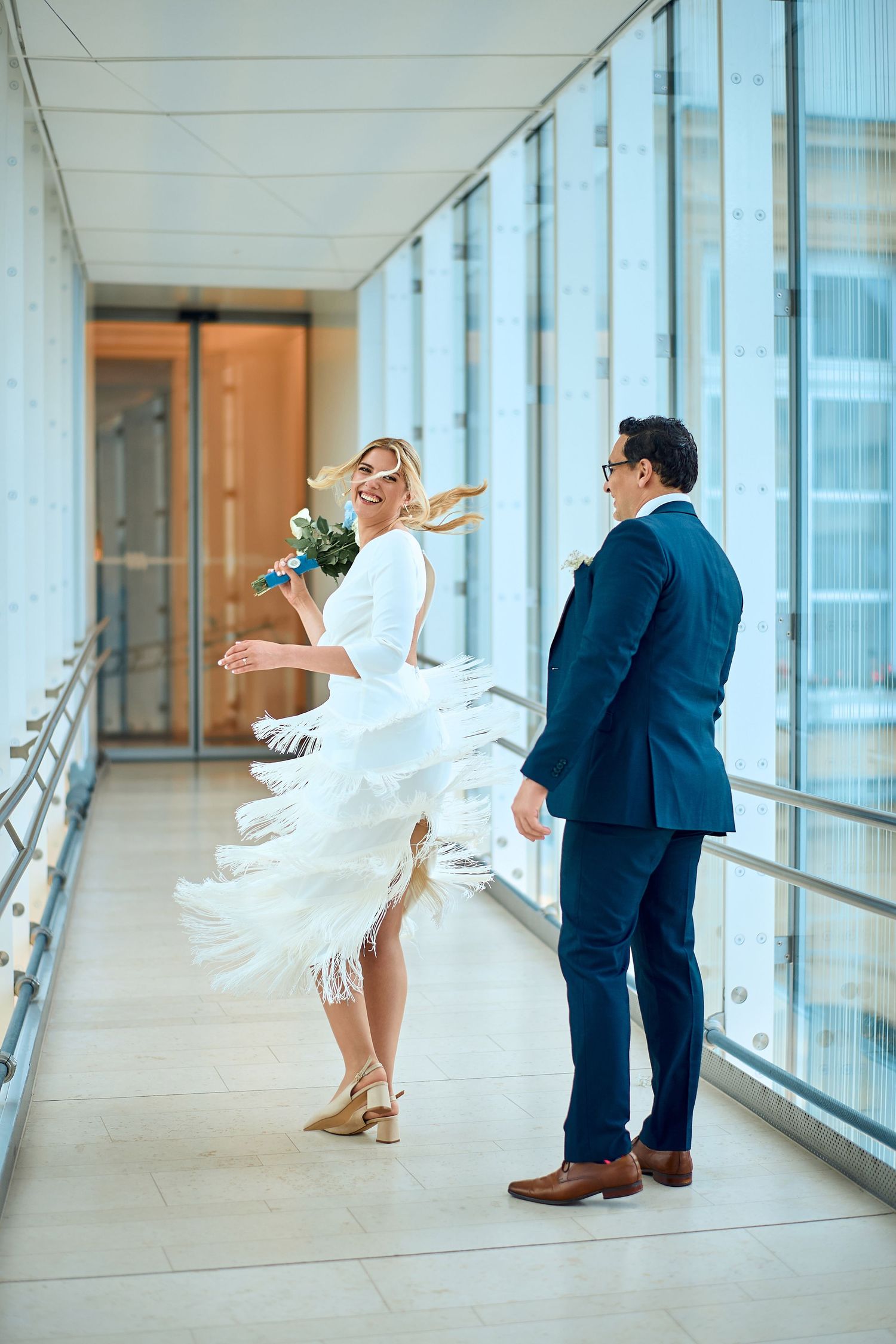 A bride twirls her dress playfully in a modern hallway while her partner watches.