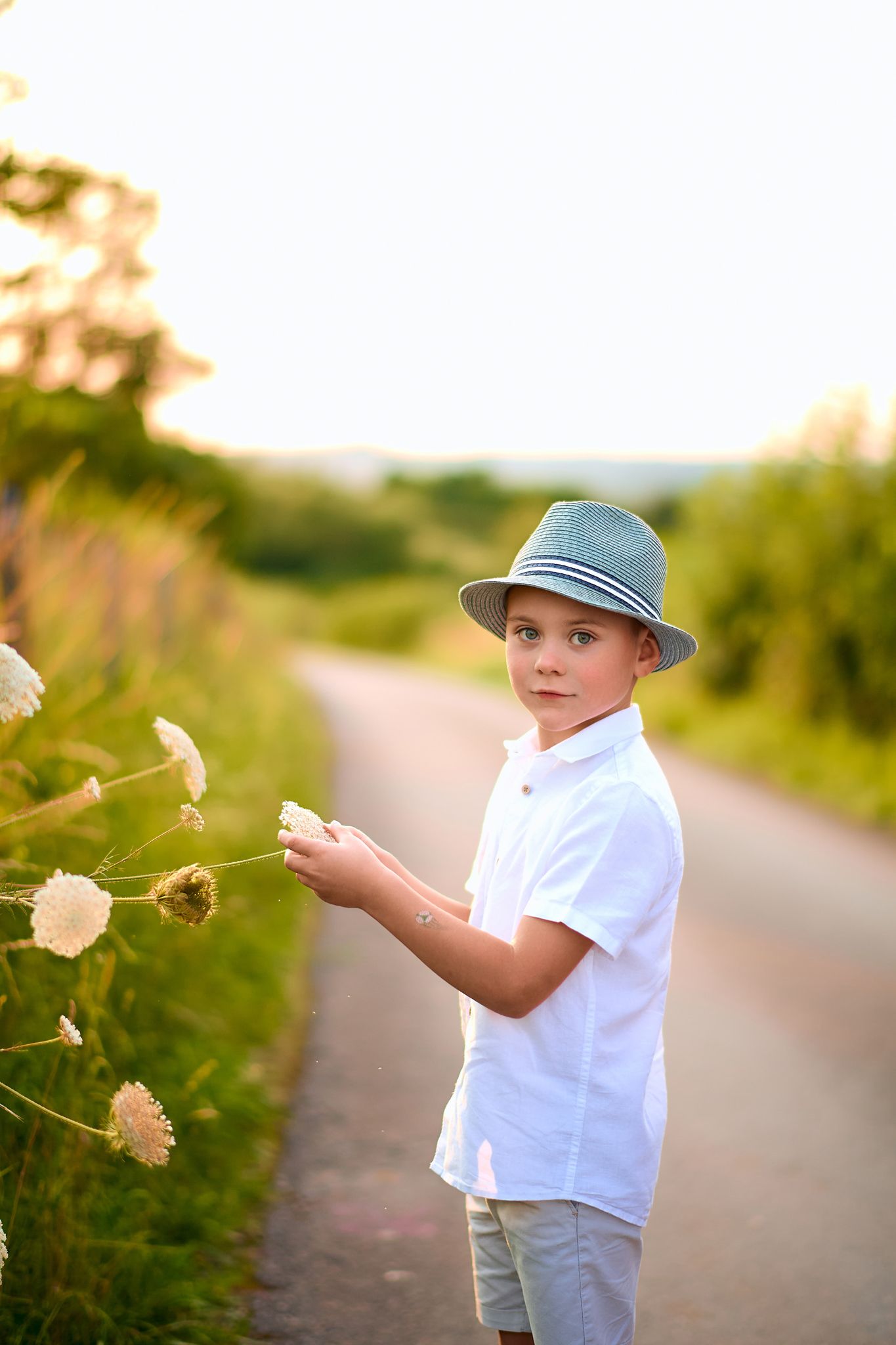 Young child in white shirt and blue hat playing with dandelions at sunset on a country path.