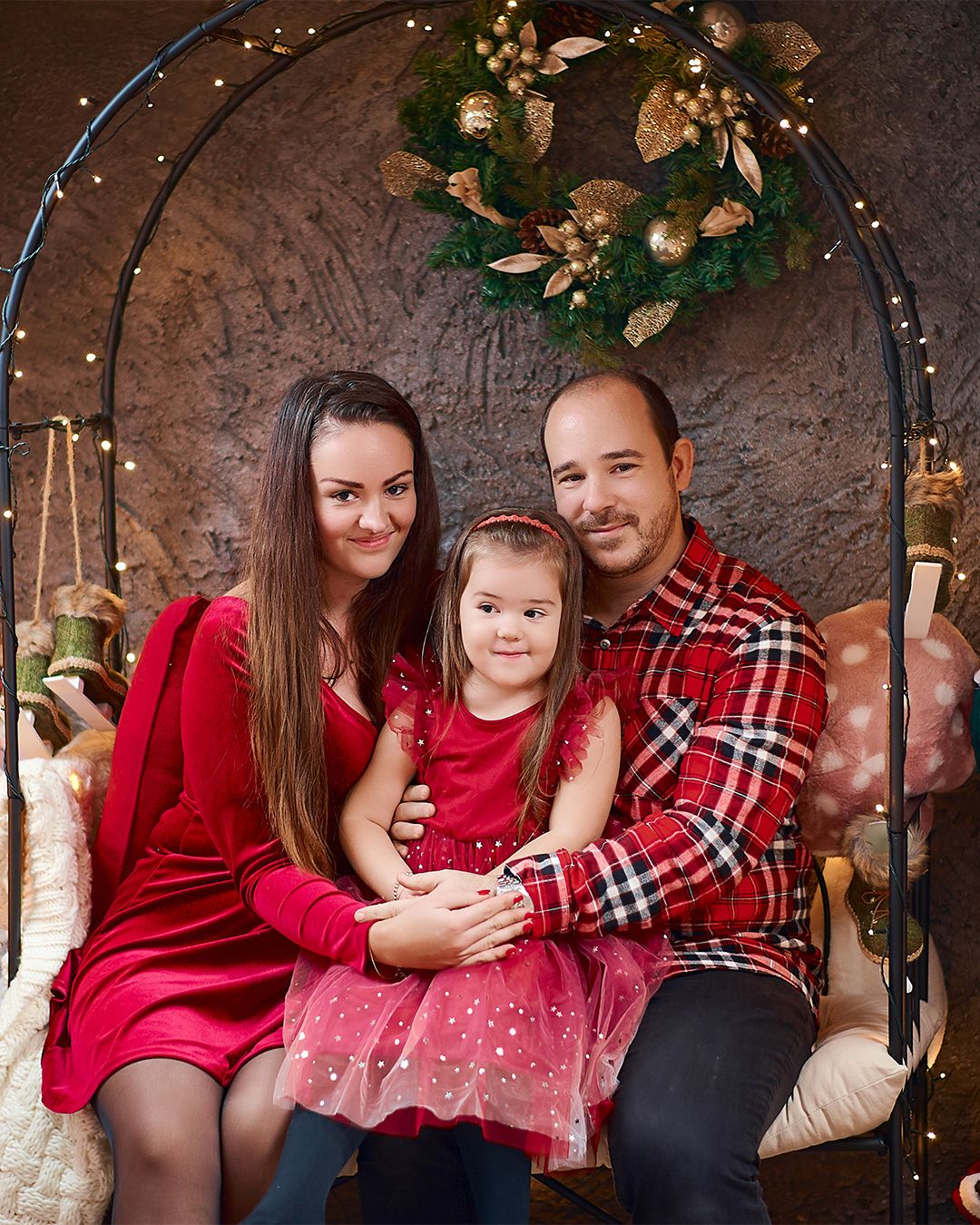Family of three in red outfits posing on a swing under a Christmas wreath with festive lights backdrop.